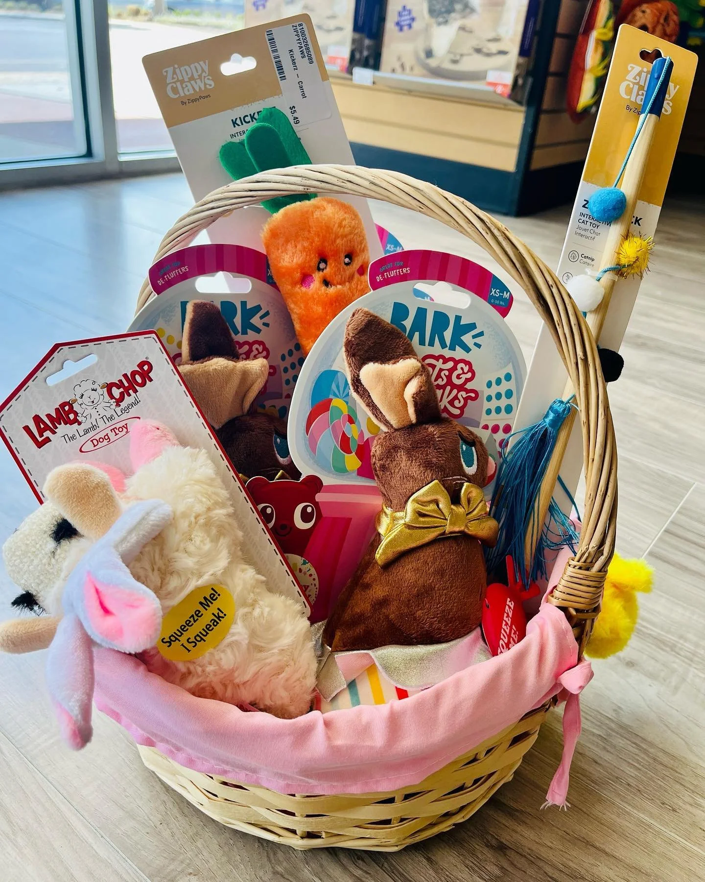 A basket filled with various plush animal toys and pet toys, including a stuffed bunny, a carrot, and dog toys, inside a store near a glass door.
