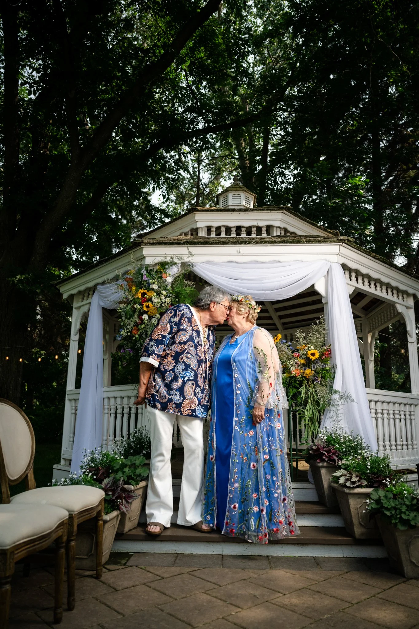 Two women, one older woman with short gray hair wearing a blue floral dress and another older woman with short gray hair in a colorful shirt and white pants, are leaning noses together in a kiss in front of a garden gazebo decorated with white fabric