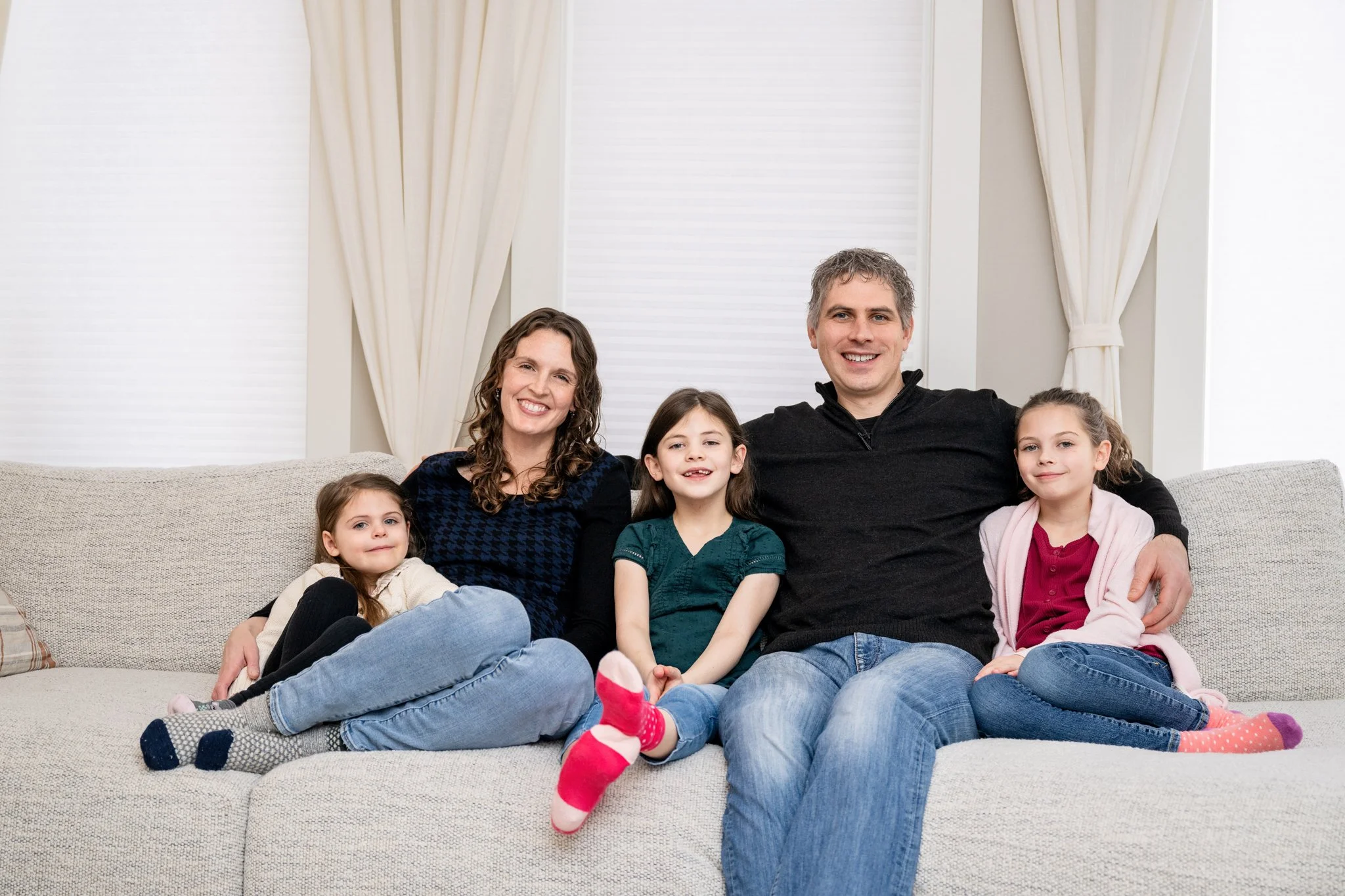 Family of five sitting on a beige couch in a living room, smiling, with white curtains behind them.