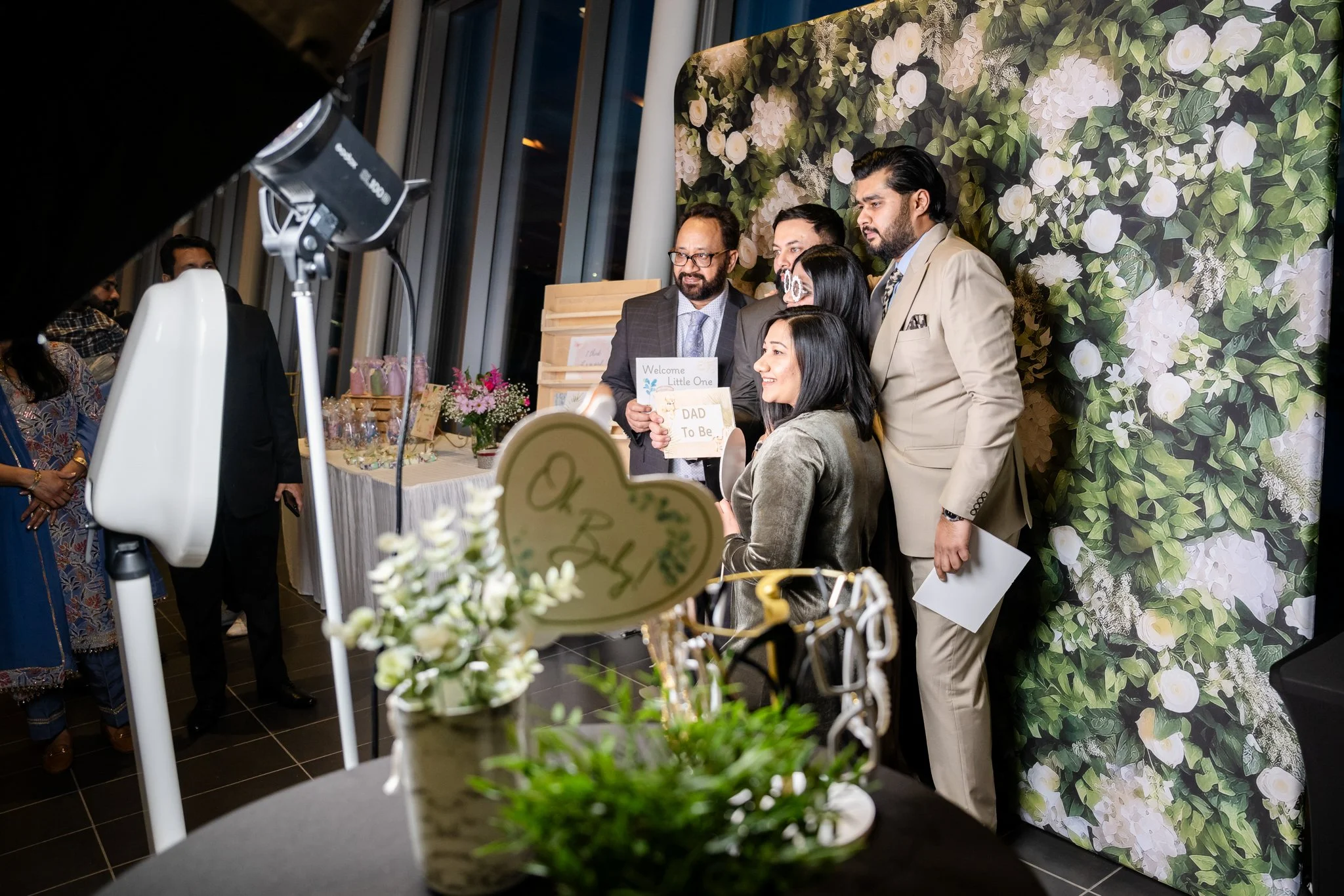 Group of people at a baby shower, with a floral backdrop, some holding signs that say "Welcome Little One" and "DAD To Be".