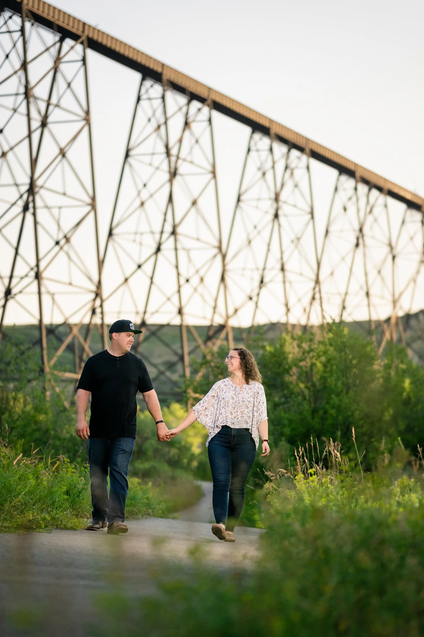A couple walking hand-in-hand on a dirt path with green trees and bushes in the background, and a large metal bridge overhead.