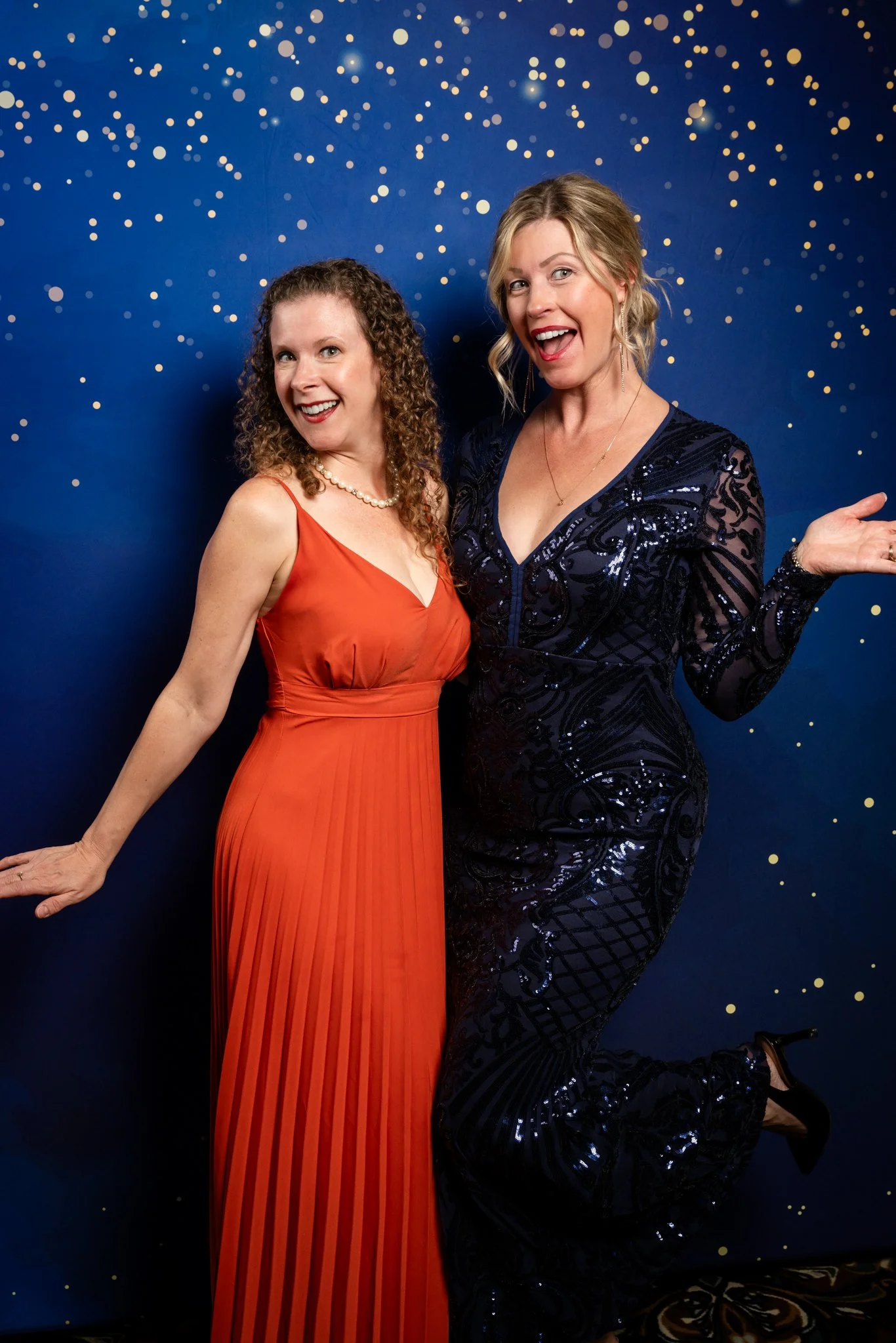 Two women in formal evening gowns smiling and posing in front of a dark blue backdrop with gold confetti or stars.