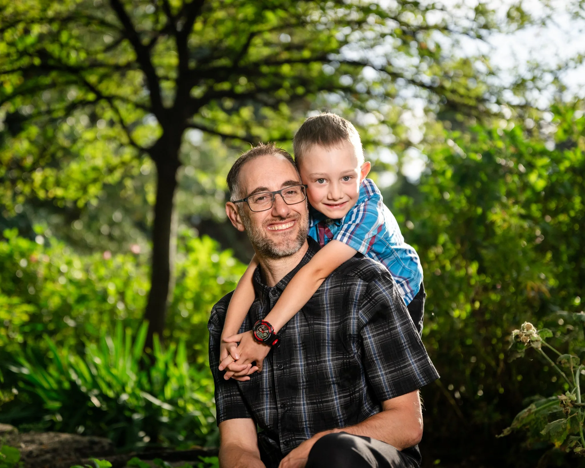 A man with glasses and a beard smiling while carrying a young boy on his shoulders in a lush green outdoor setting.