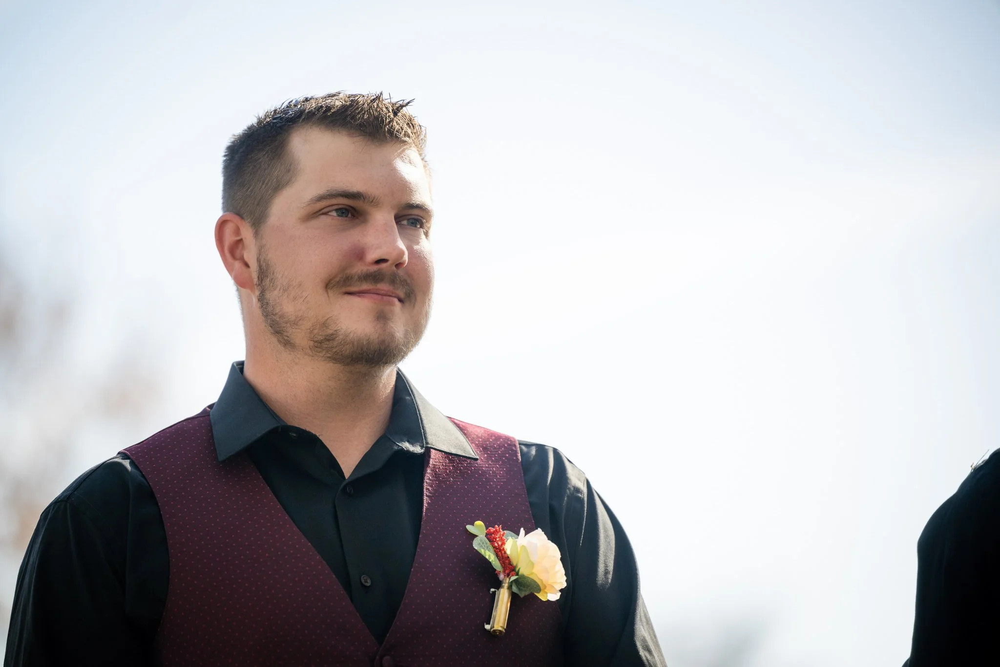 A young man with short brown hair, a beard, and wearing a black shirt with a maroon vest and a boutonniere, standing outdoors with a bright sky in the background.