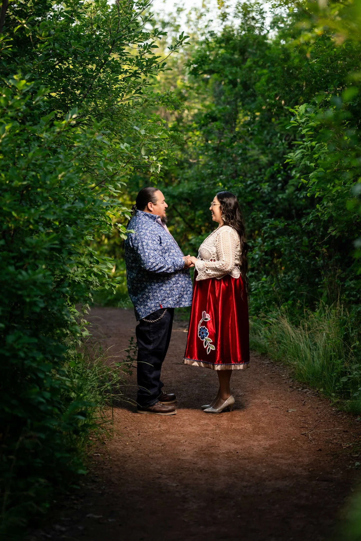Two women standing face-to-face on a dirt path in a green forest, holding hands and smiling at each other, dressed in traditional and modern clothing.