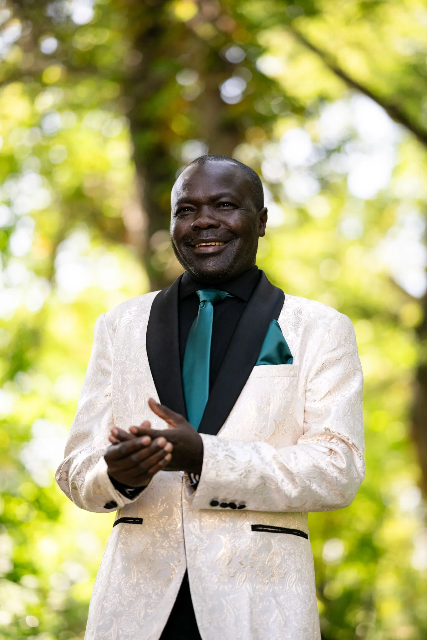 A man in a white suit with black and teal accents smiling outdoors with green trees in the background.