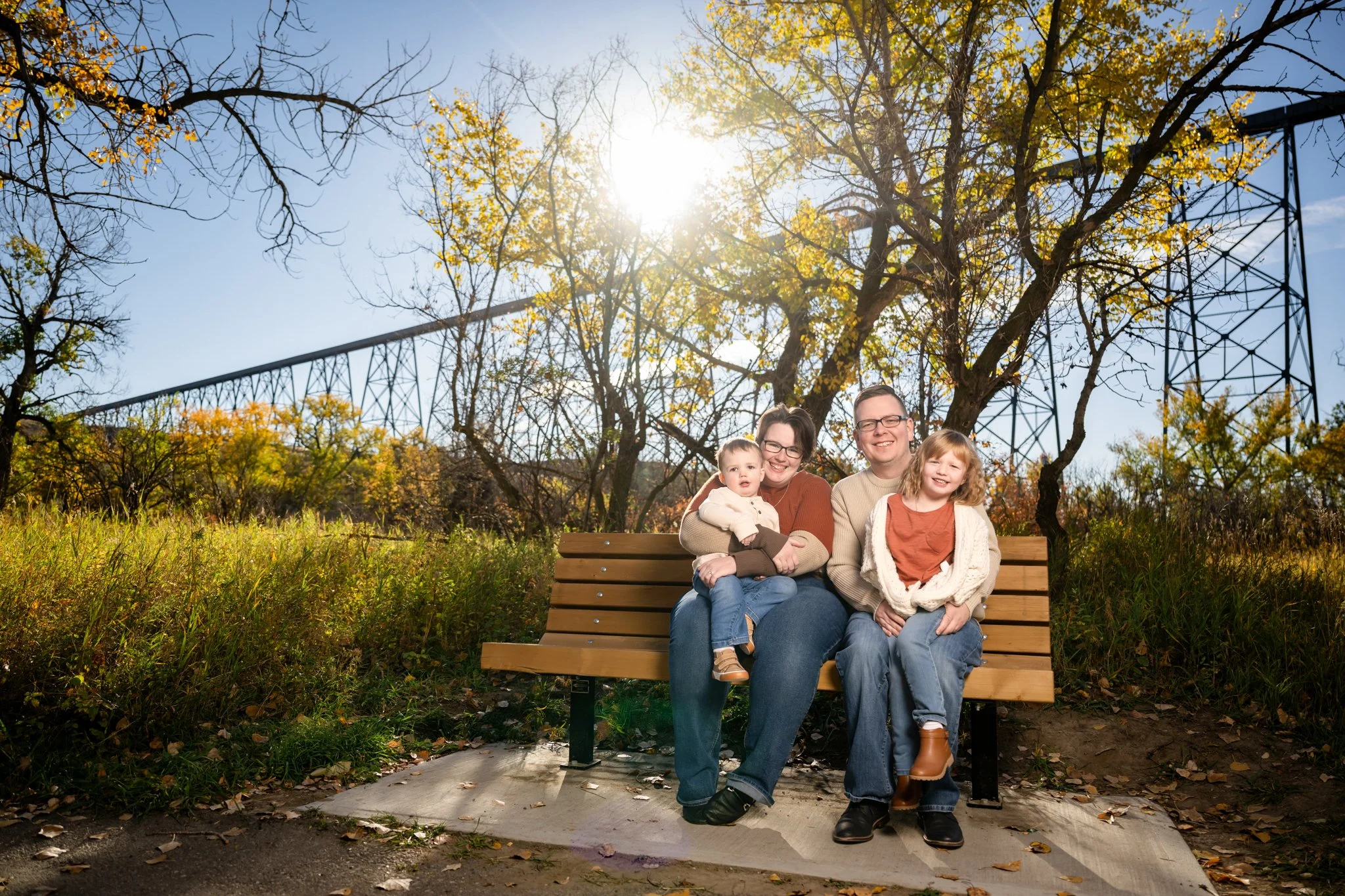 Family of four sitting on a park bench outdoors during autumn, with trees and a clear sky in the background.