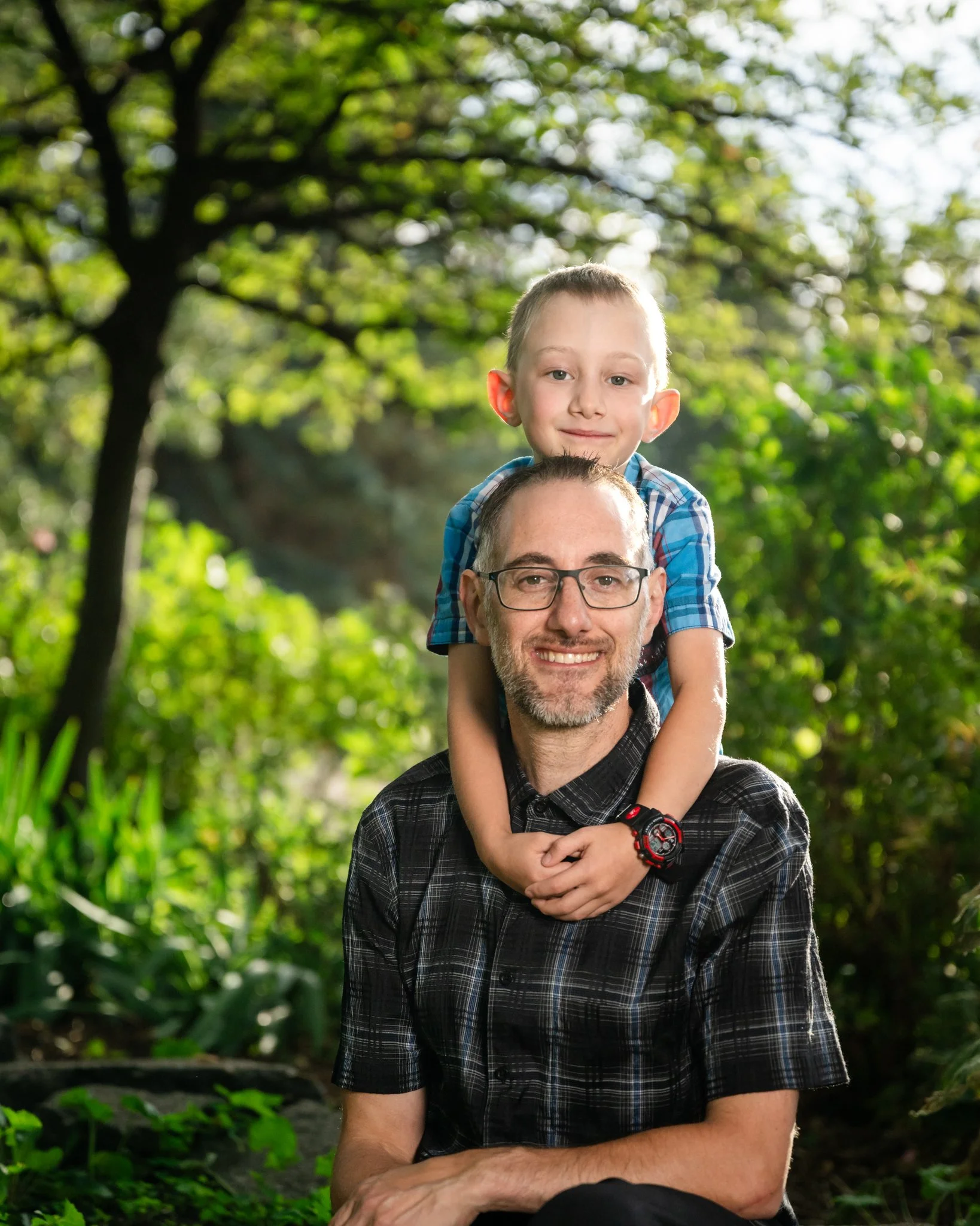 A man with glasses and a beard sitting outdoors in a lush green forest, smiling, with a young boy on his shoulders, also smiling.