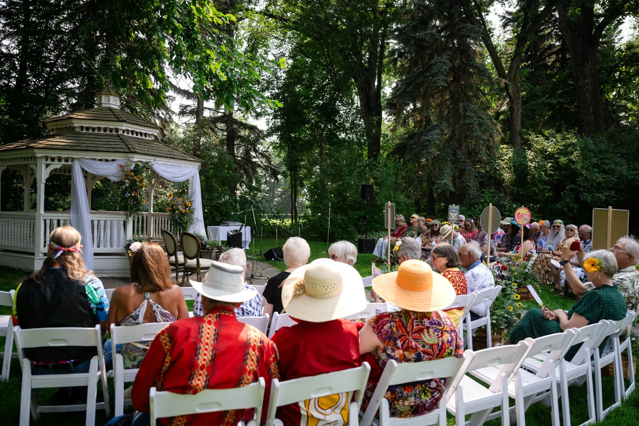 People gathered outdoors for a wedding ceremony in a garden, with a gazebo decorated with flowers and white drapes, surrounded by trees.