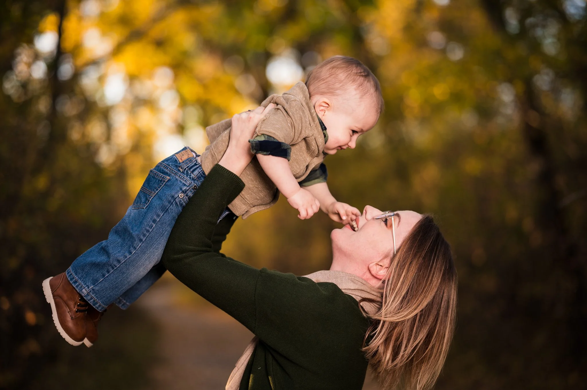 A woman with glasses and brown hair lifting a smiling young boy with blonde hair, wearing a brown shirt and jeans, in an outdoor setting with fall foliage.