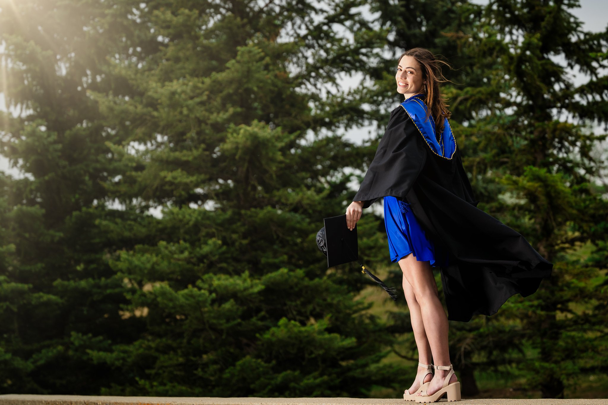 graduate standing in blue and black grad gown at sunset
