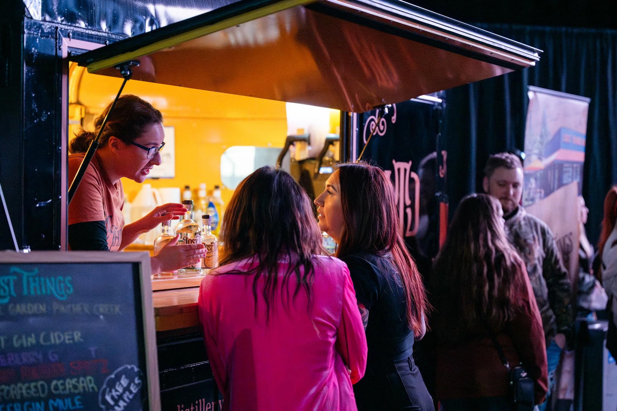 A bartender serving alcohol to two women at an outdoor booth with a black sign and neon lettering, with a line of people waiting.