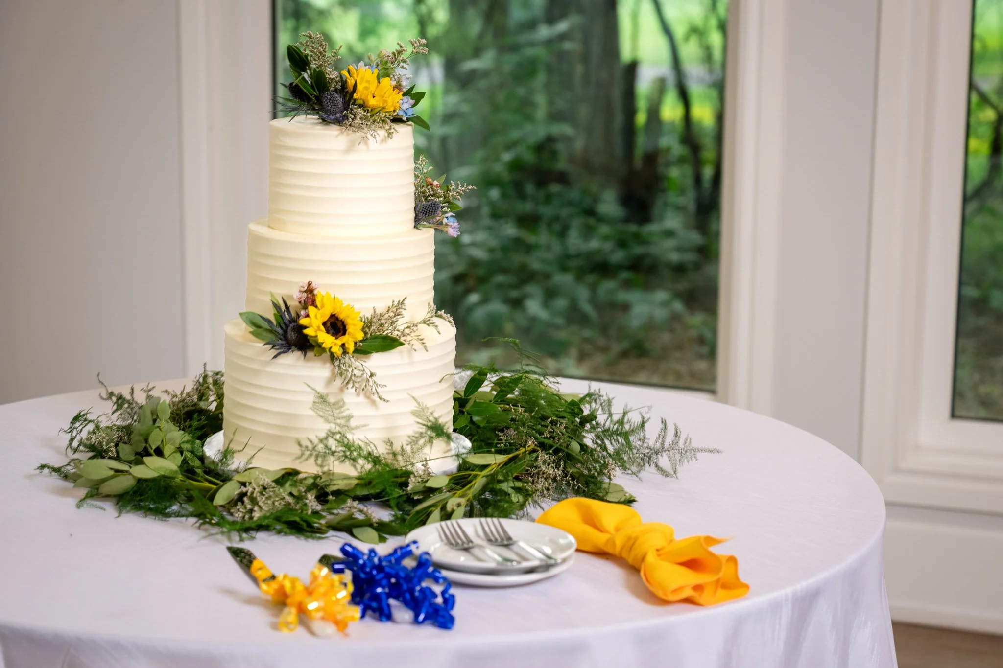 Three-tier white wedding cake decorated with sunflowers, greenery, and small flowers, placed on a table with a white tablecloth, yellow napkin, and small plates and forks, with greenery surrounding the base, near a window with a view of a green outdo