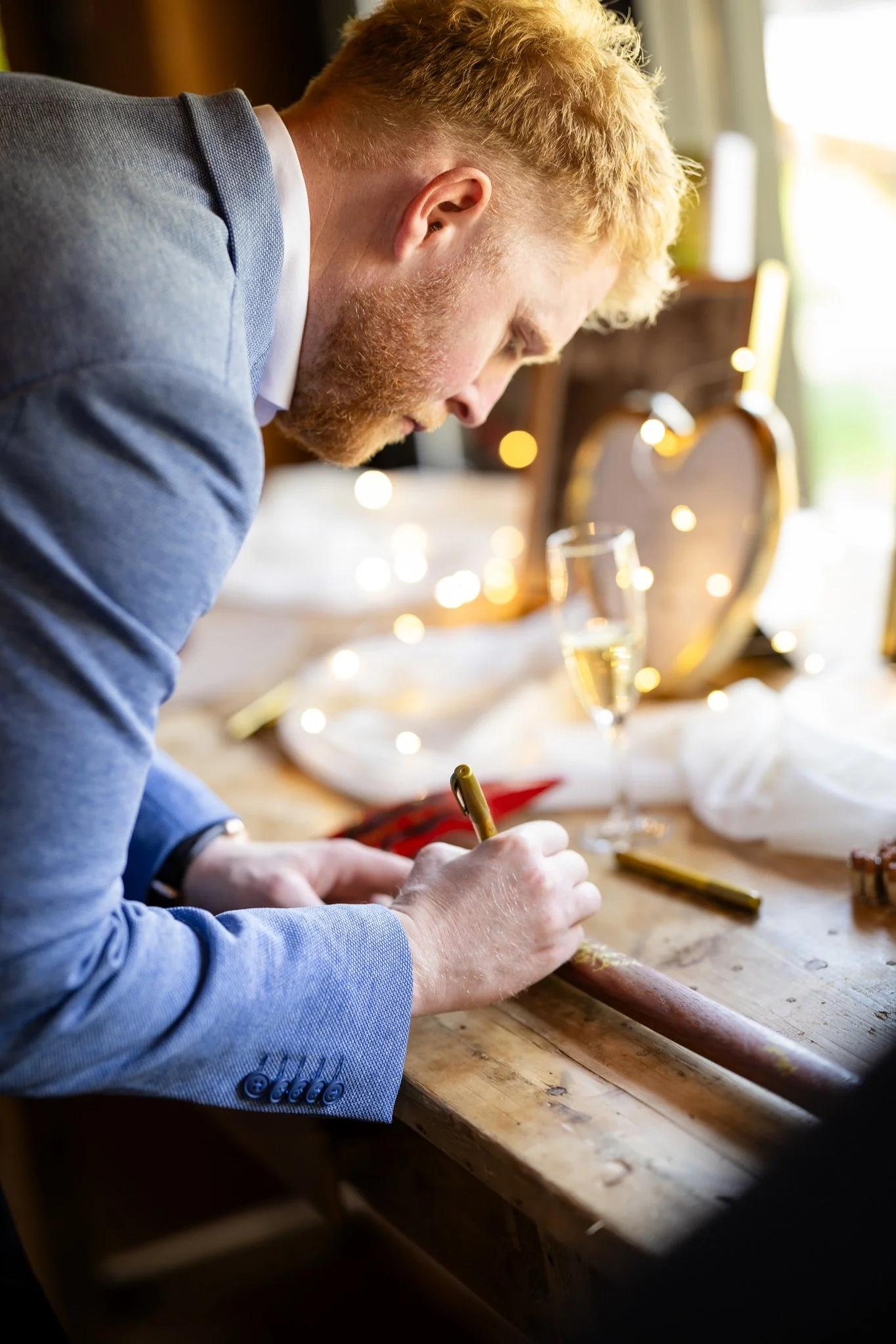A man in a light blue suit is writing on a wooden surface, with a glass of champagne and a heart-shaped photo frame in the background, illuminated by warm string lights.