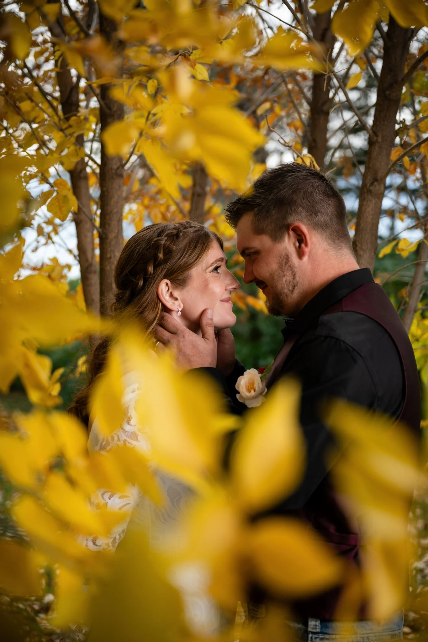 A couple gazing into each other's eyes with their faces close, surrounded by autumn leaves and trees.