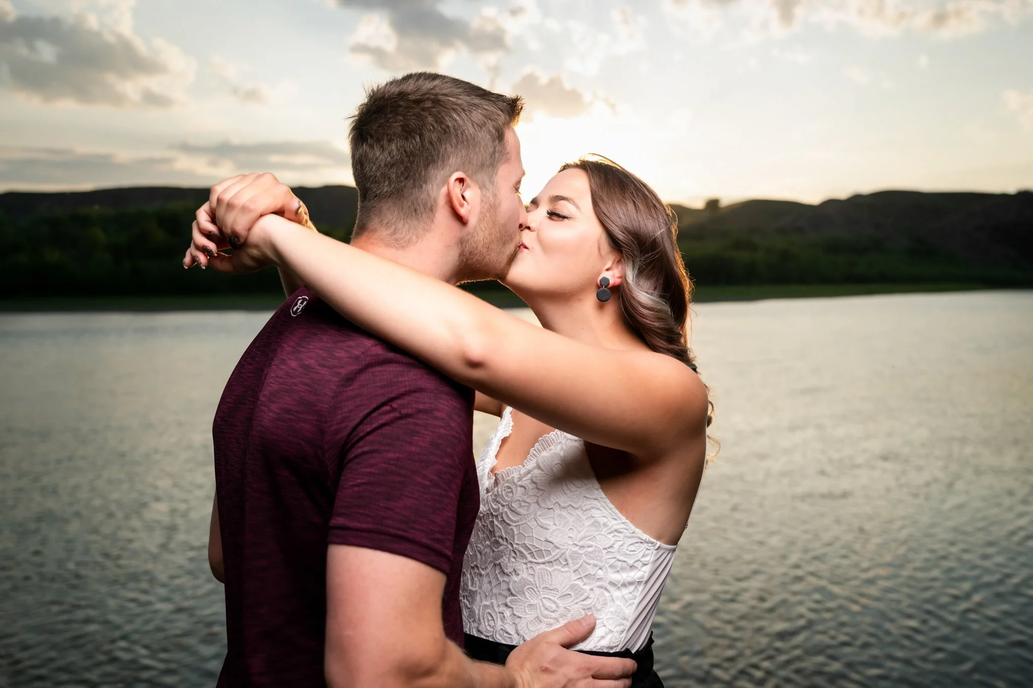 A couple kissing by a lake during sunset. The woman has her arms around the man's neck, and the man is holding her waist. The background features a calm lake and a hilly landscape.