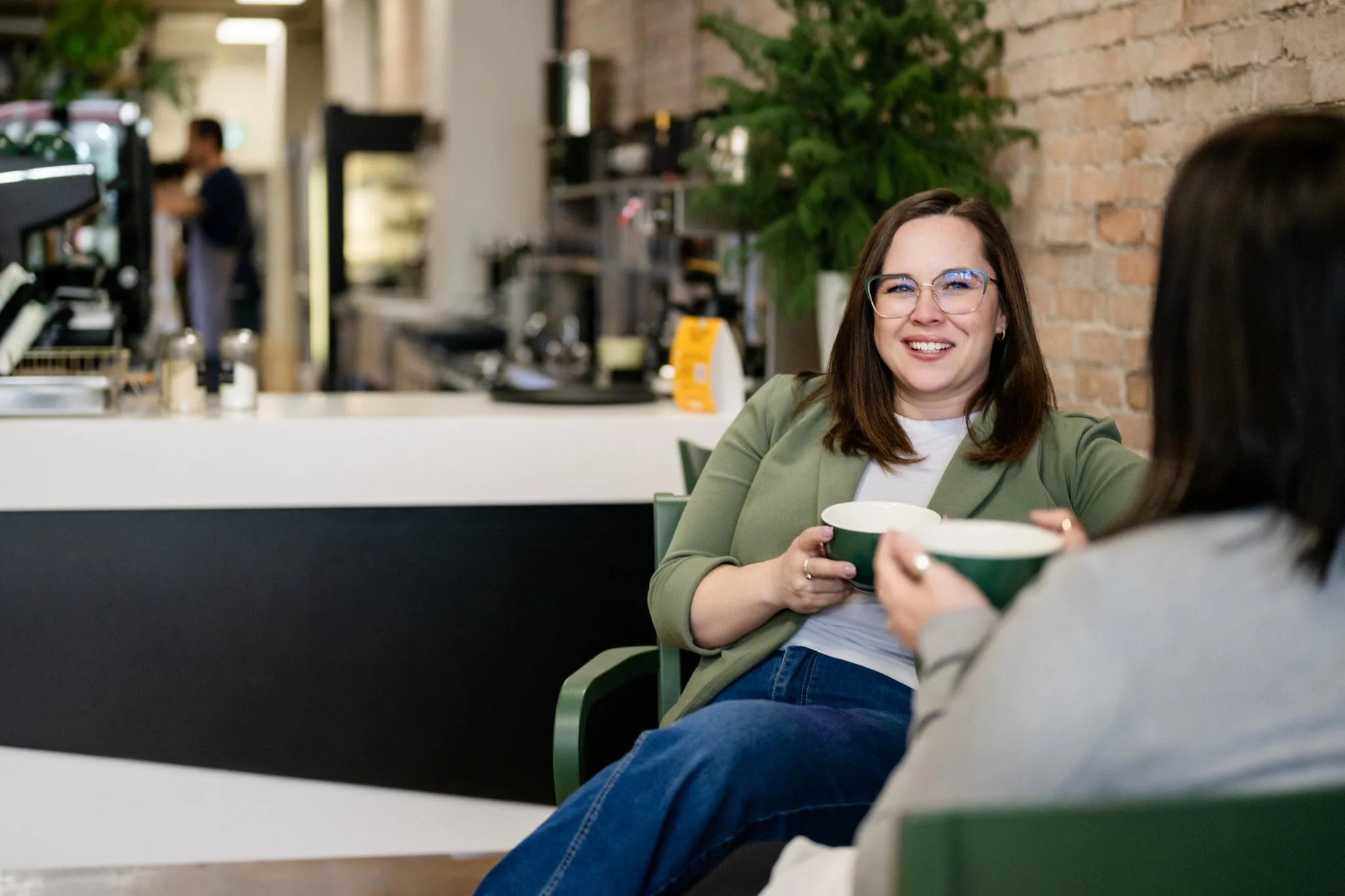 Two women sitting in a cafe, one with glasses smiling and holding a green bowl, the other with dark hair also holding a green bowl, with a cafe counter and kitchen in the background.