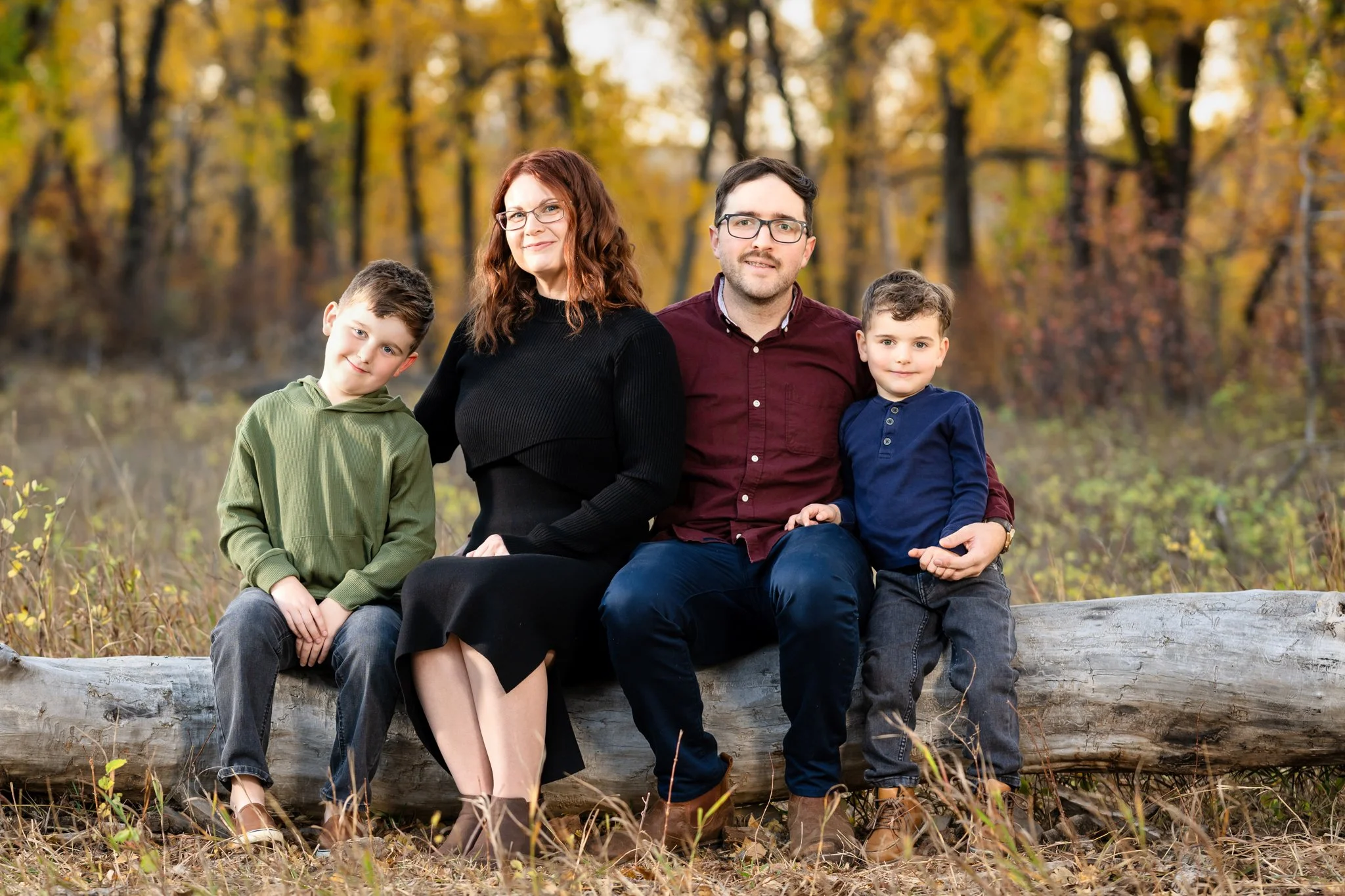 A family of four sitting on a log in a fall forest, with trees and autumn leaves in the background.