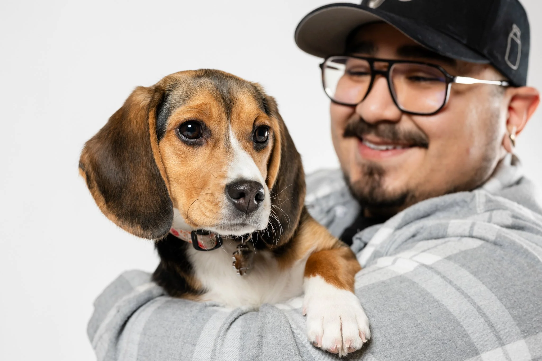 A man wearing glasses, a hat, and a gray hoodie is smiling and holding a beagle puppy with brown, black, and white fur against a plain white background.