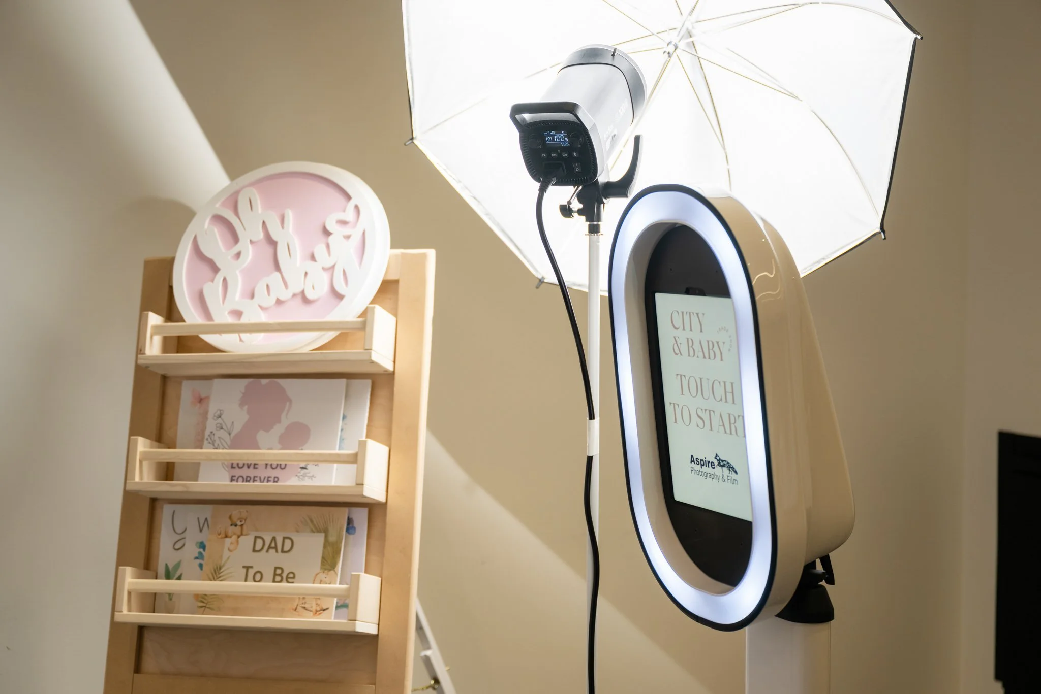 Photo booth setup with a ring light, umbrella light, and a touchscreen sign that reads 'City & Baby Touch to Start,' along with greeting cards and artwork for a baby shower or celebration.