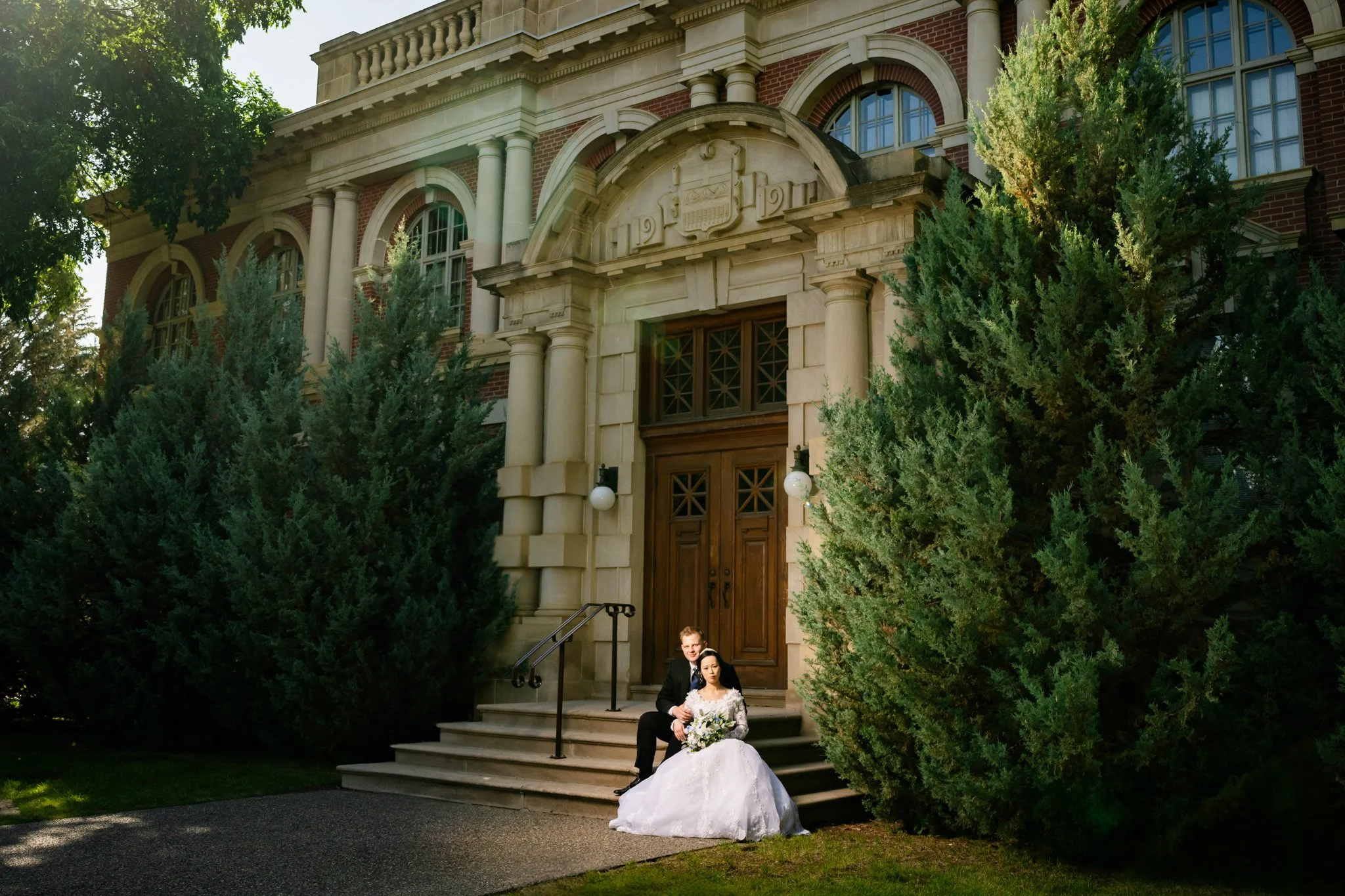 A bride and groom sitting on the steps of a historic brick building with large wooden doors, surrounded by greenery and tall bushes.