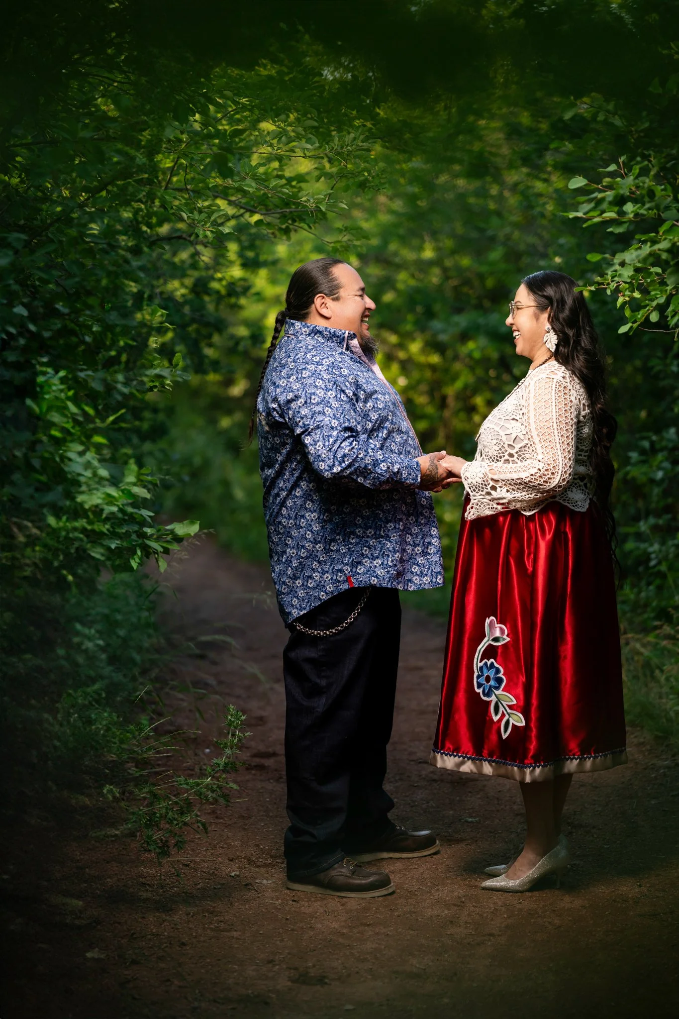 A couple holding hands and smiling at each other on a dirt path surrounded by green foliage in a forest.