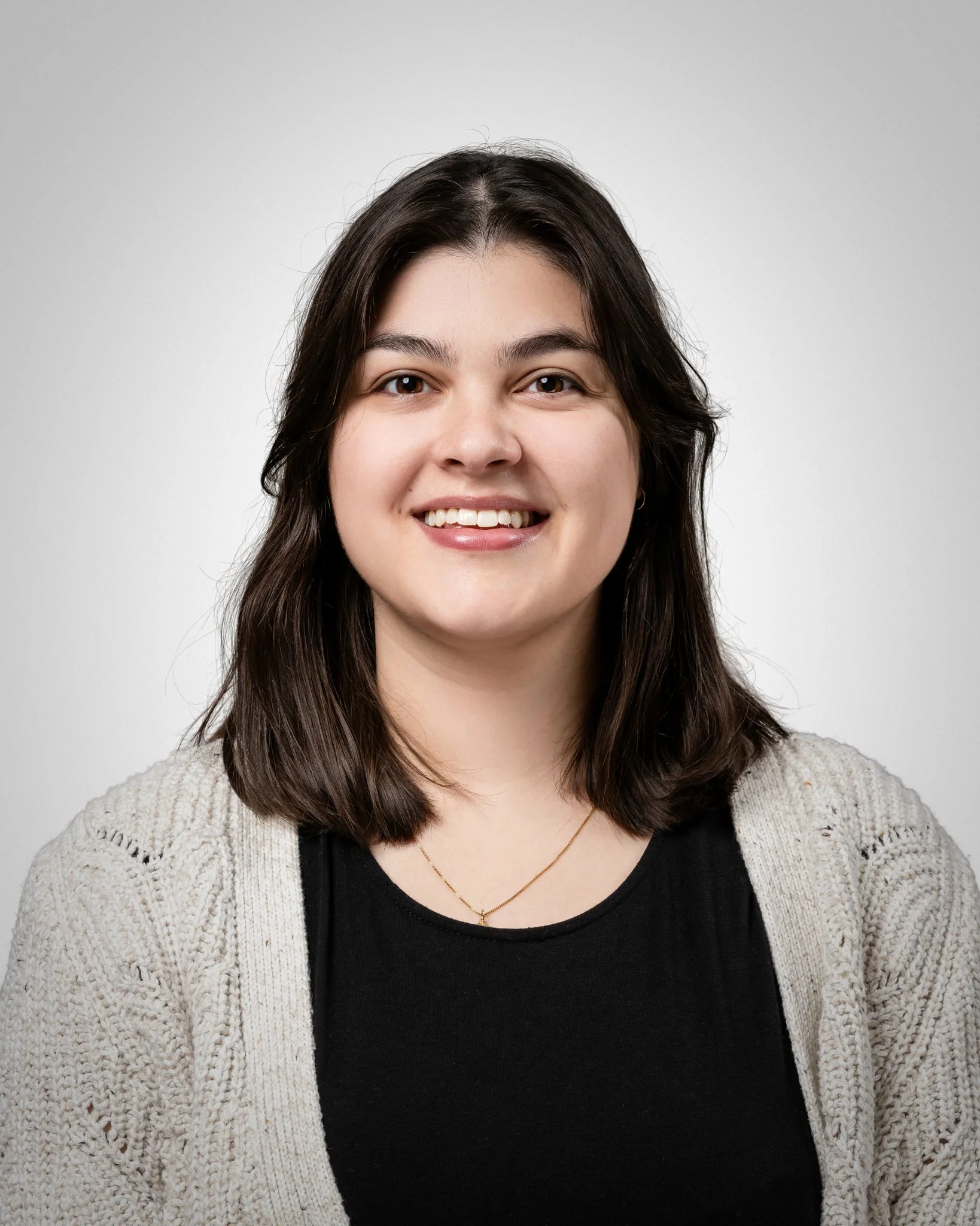 A young woman with shoulder-length dark brown hair, smiling, wearing a black top, a cream-colored cardigan, a gold necklace, against a plain light background.