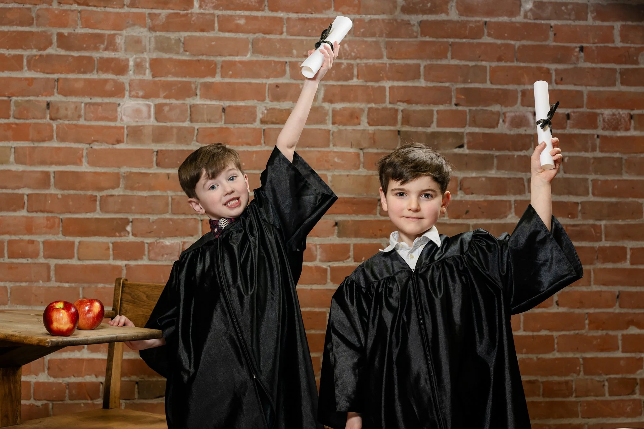 Two young boys wearing black graduation gowns and holding rolled-up diplomas in front of a brick wall. One boy is smiling, and the other looks content. Apples rest on a wooden table nearby.