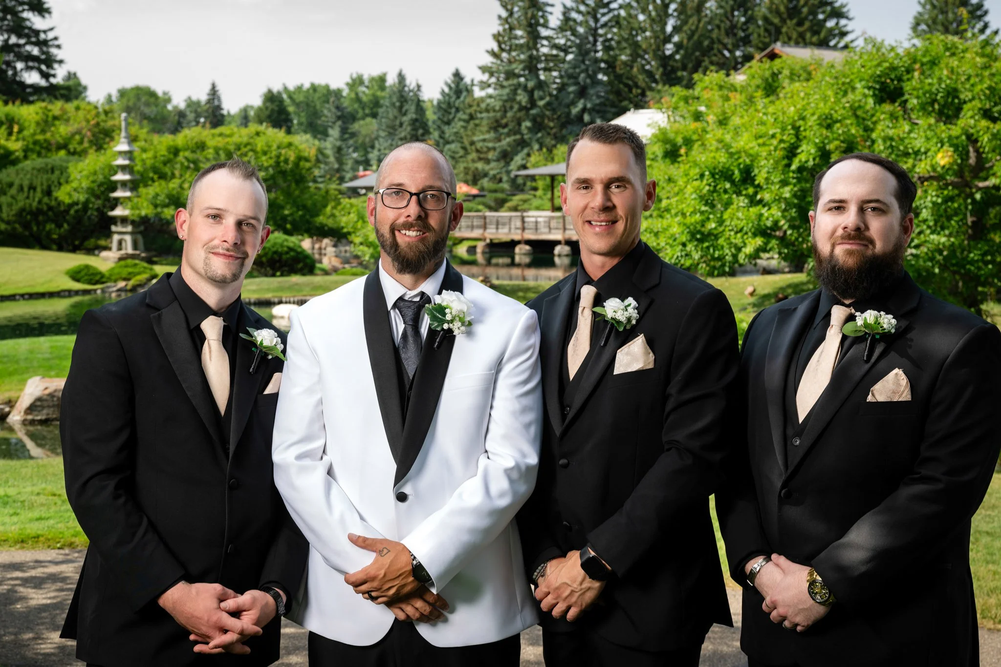 Four men dressed in tuxedos, with the groom in a white tuxedo and the groomsmen in black tuxedos, standing outdoors in front of a lush green landscape with trees, a pond, and a bridge.