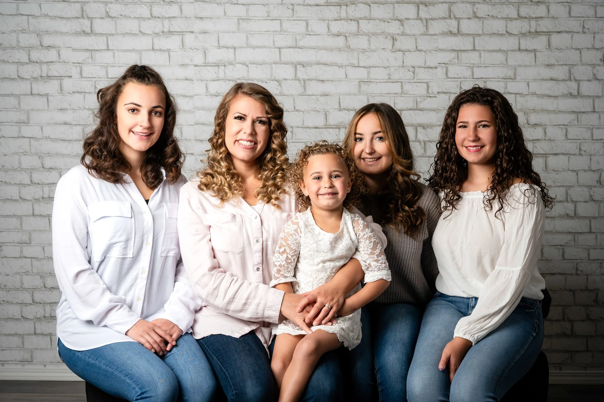 Six women, including a young girl, sitting and smiling against a white brick wall.