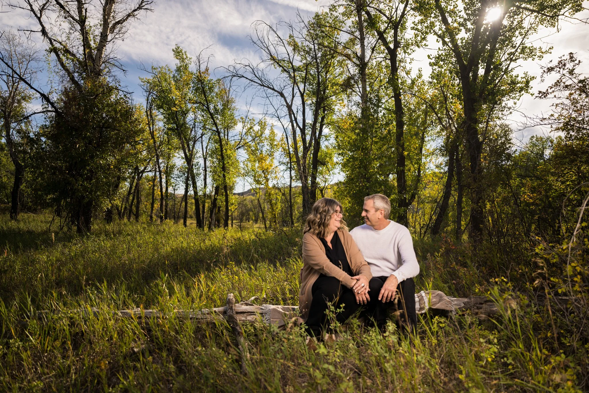 A middle-aged couple sitting on a fallen log in a forest clearing, enjoying each other's company on a sunny day with green trees and blue sky in the background.