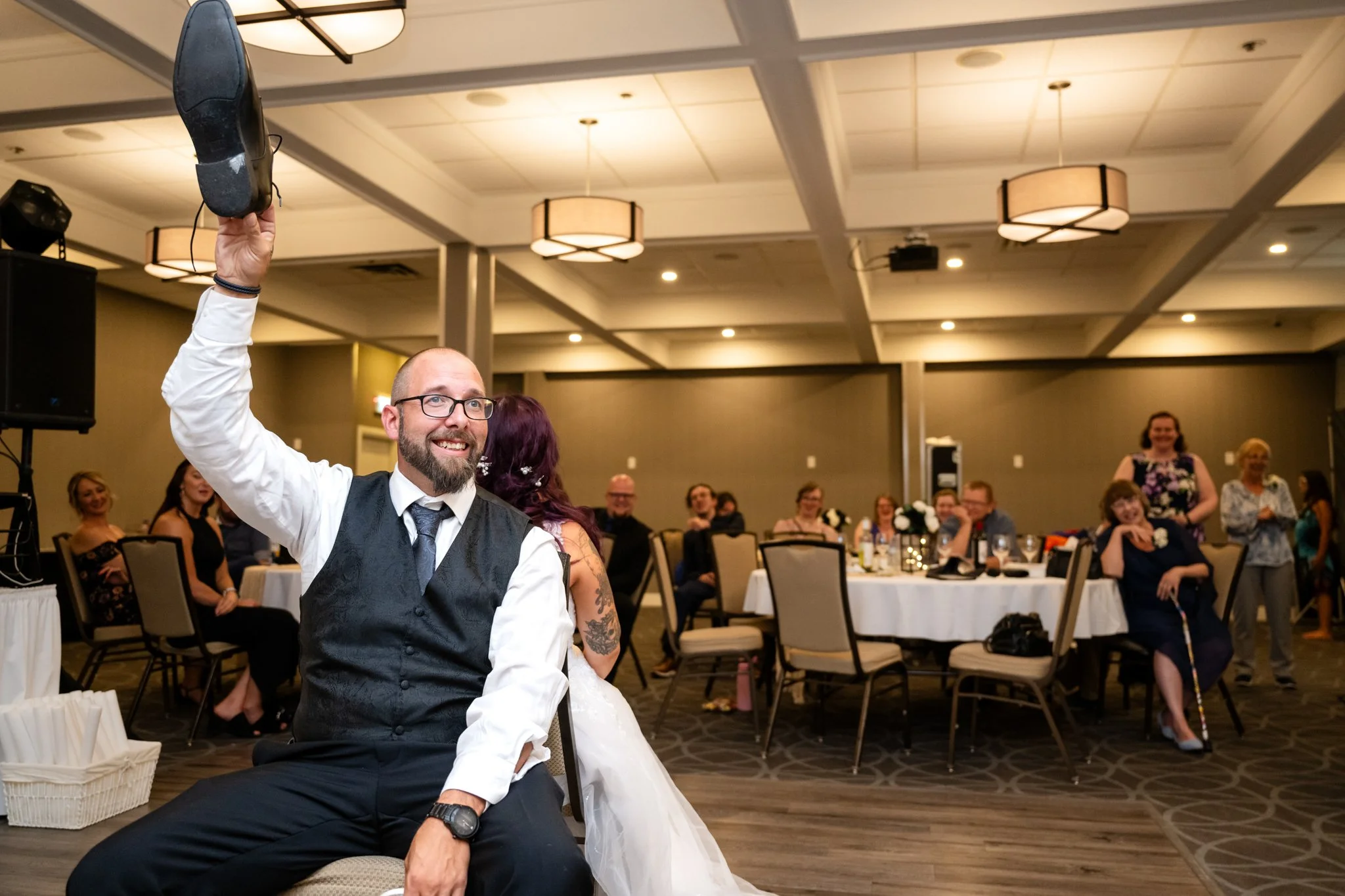 A man in formal attire with glasses holding a shoe high in the air at a wedding reception while guests look on and laugh.