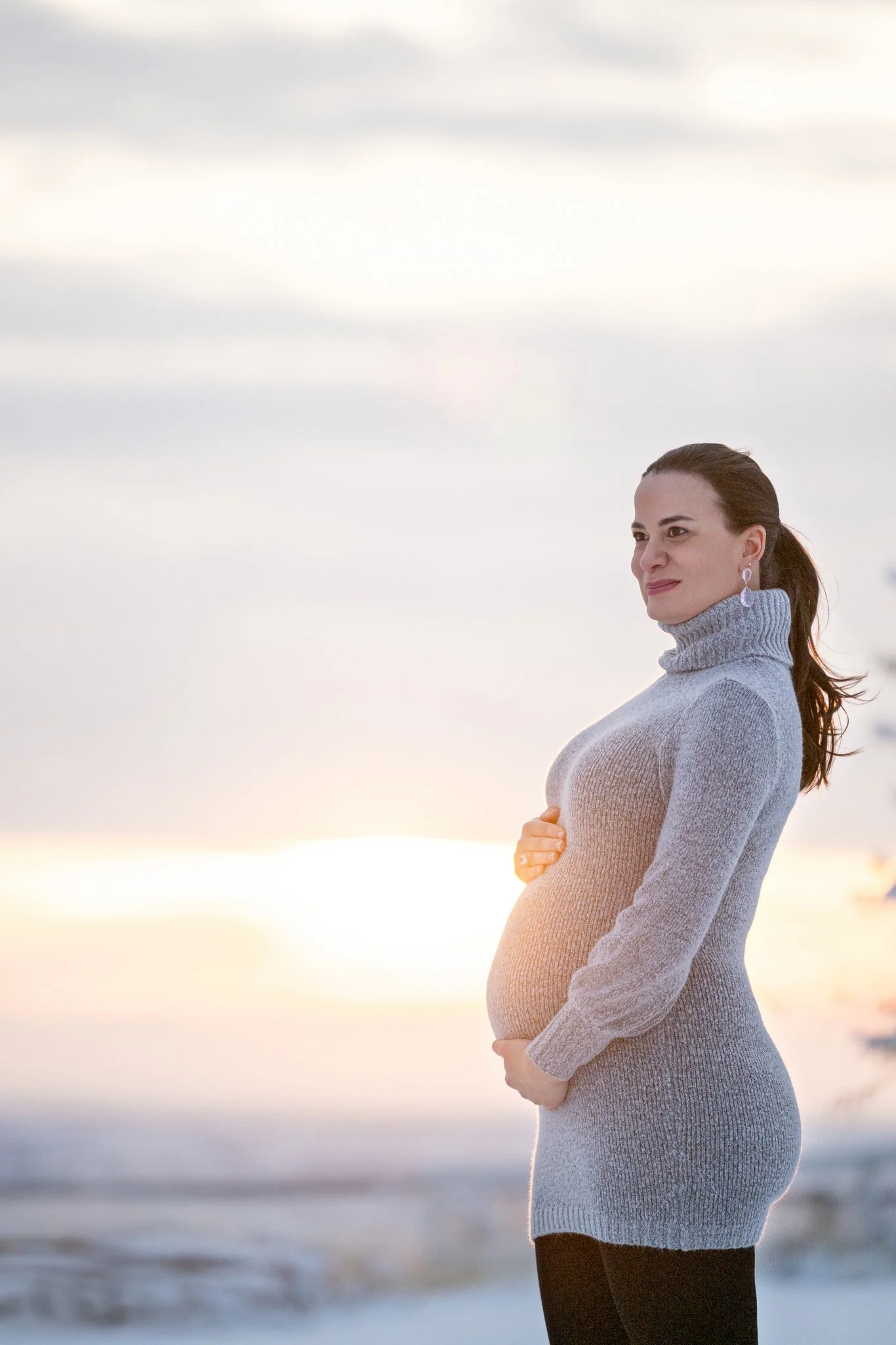 A pregnant woman in a gray turtleneck sweater standing outdoors at sunset or sunrise, with her hand on her belly, looking contemplative.