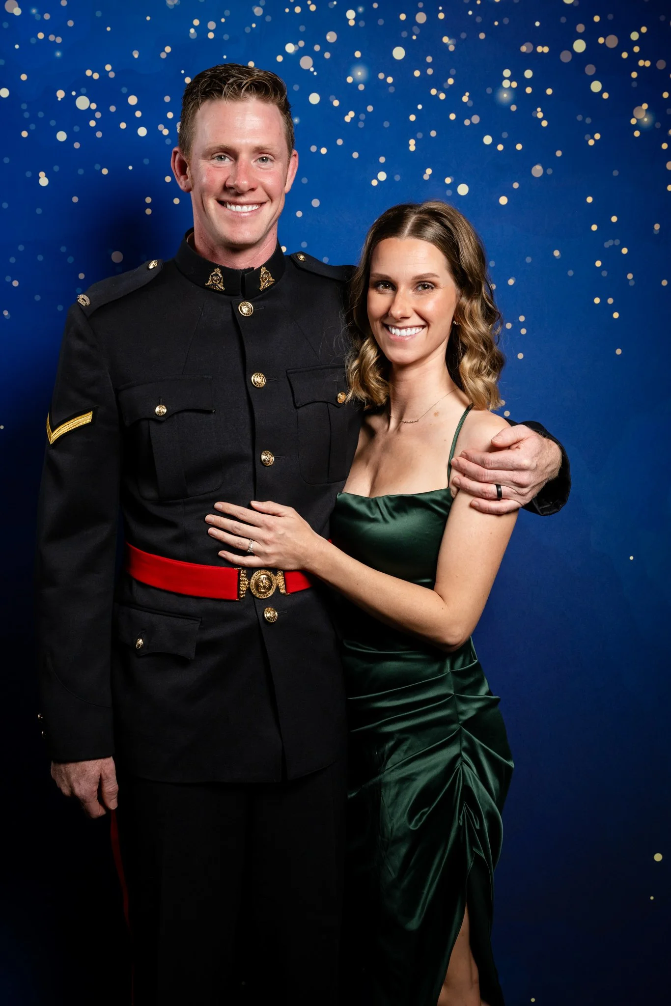 A man in a United States Marine Corps dress uniform with a woman in a green satin dress, both smiling and embracing each other, standing in front of a blue background with gold confetti-like dots.