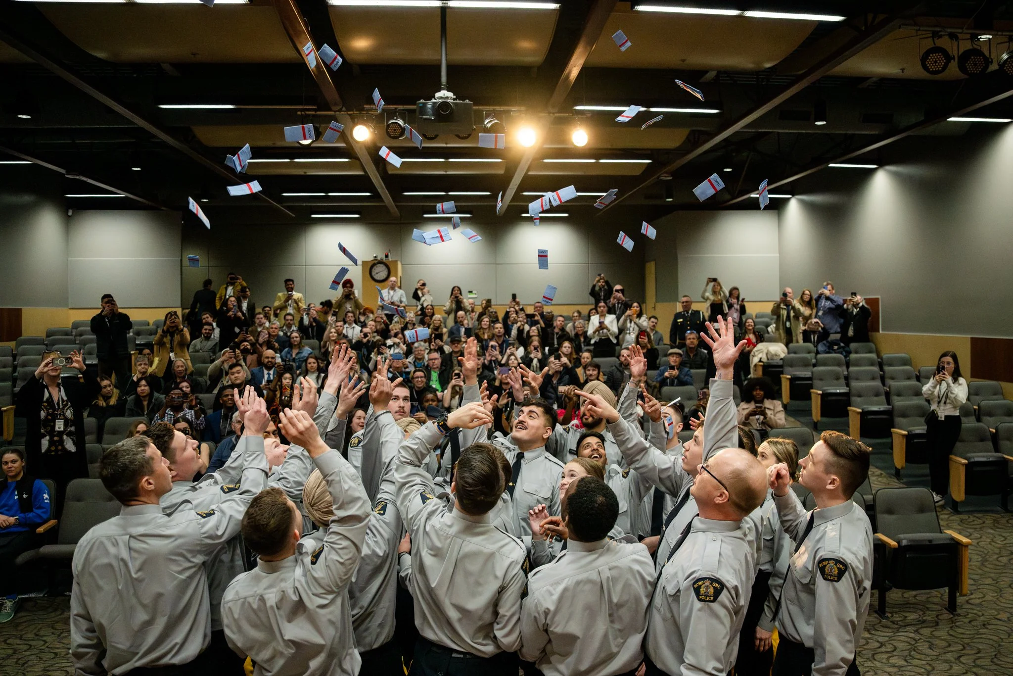 Group of officers in uniform celebrating and throwing papers into the air during a ceremony, with a large audience seated in the background.