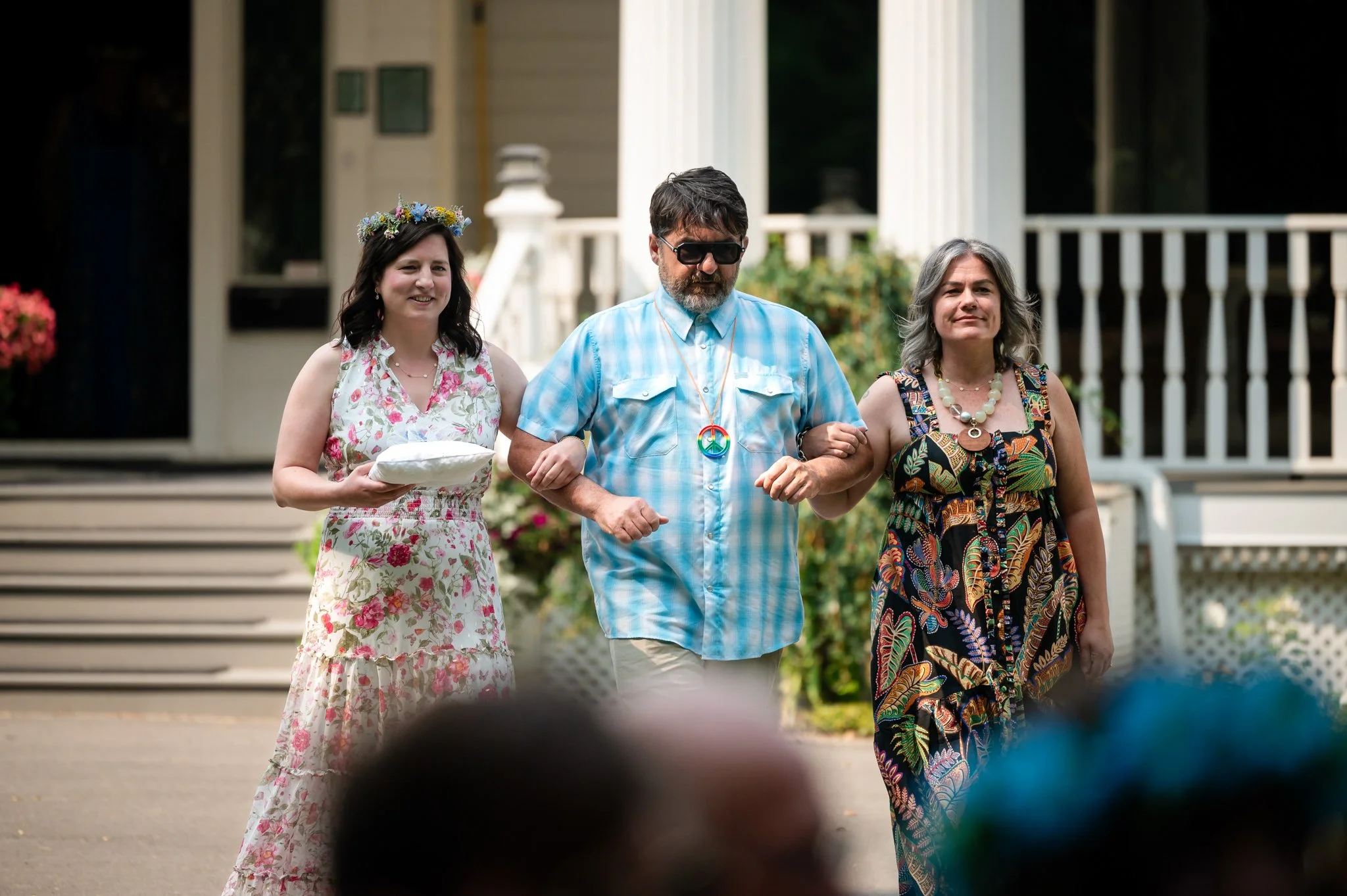 A man with sunglasses and a peace sign necklace is being walked down the aisle by two women at an outdoor wedding ceremony.