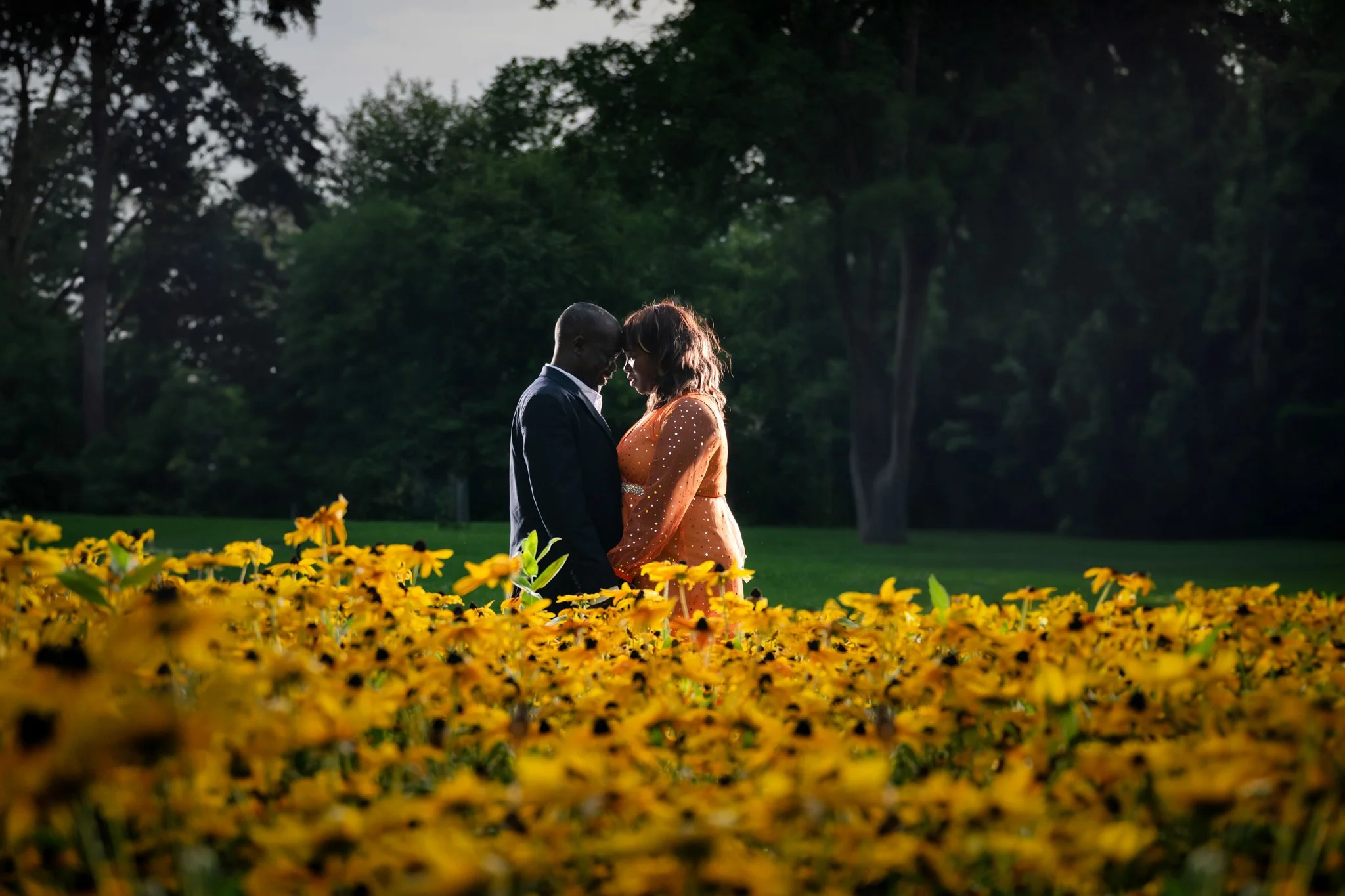 A couple standing close together in a park surrounded by yellow flowers, with trees and a cloudy sky in the background.