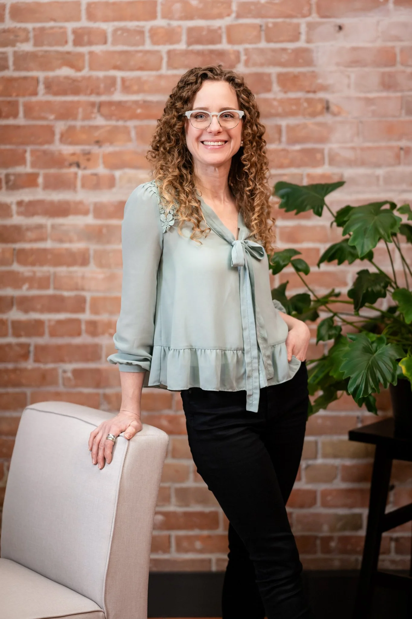 A woman with curly hair and glasses standing next to a chair, smiling, with a brick wall and a large green plant in the background.