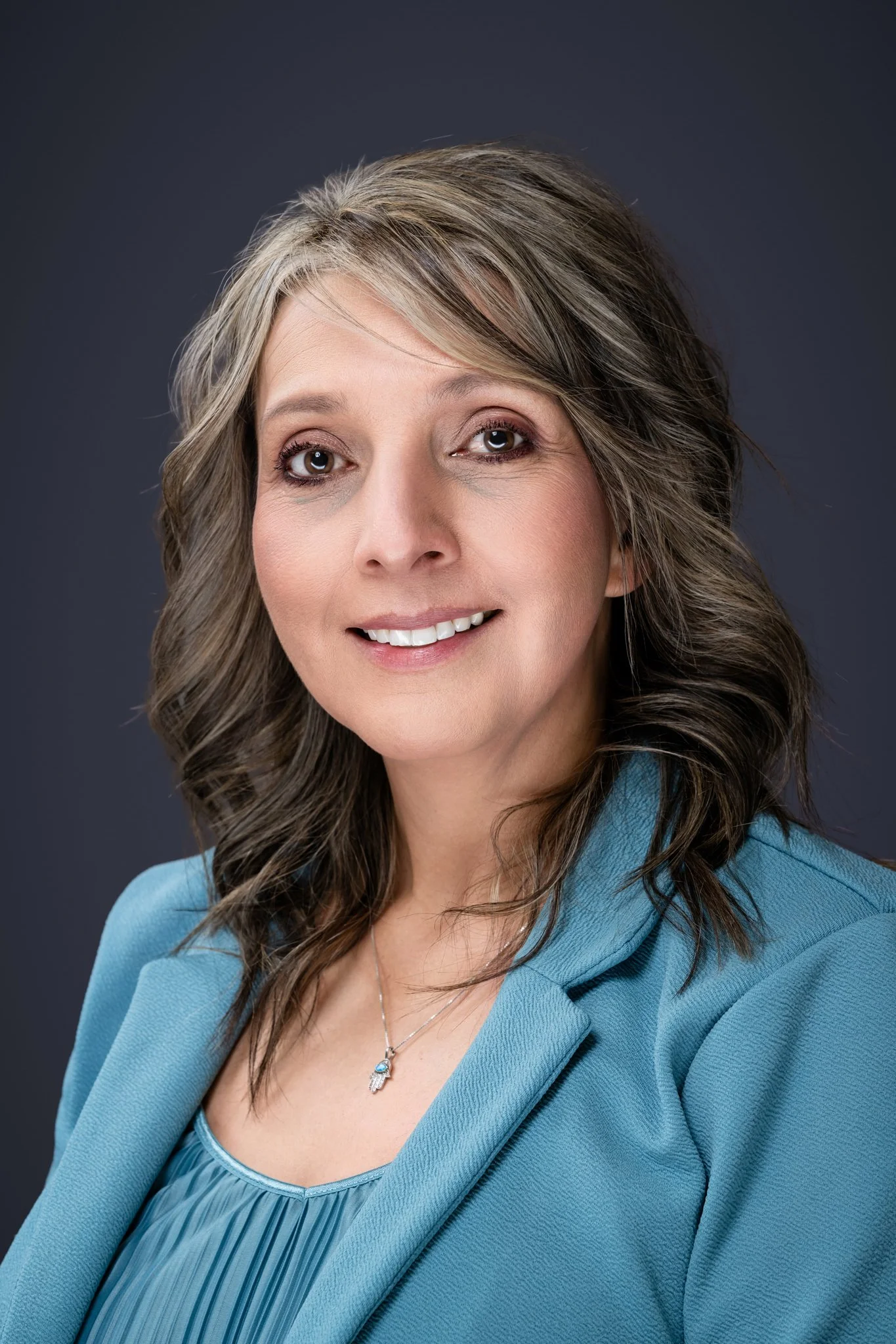 A woman with shoulder-length wavy hair wearing a blue blazer and a necklace, smiling against a dark background.