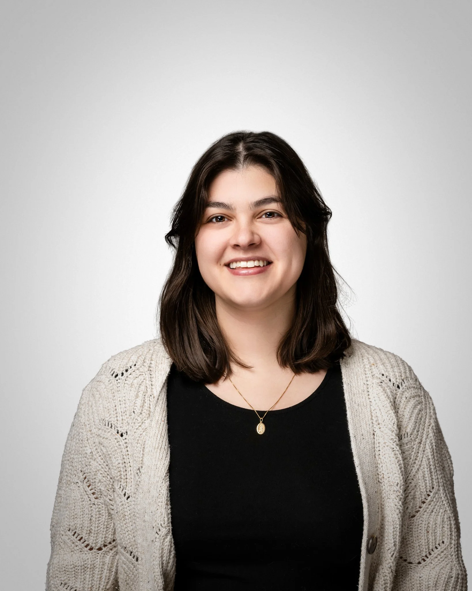 Portrait of a young woman with brown hair, smiling, wearing a black top, a beige knit cardigan, and a gold necklace with a pendant, against a plain light gray background.