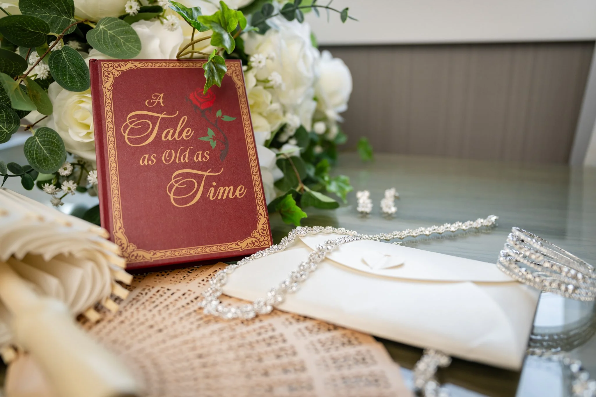 A floral arrangement of white roses and greenery, with a book titled "A Tale as Old as Time," paired with elegant jewelry including a pearl bracelet and a rhinestone necklace, and a white napkin and envelope on a table.