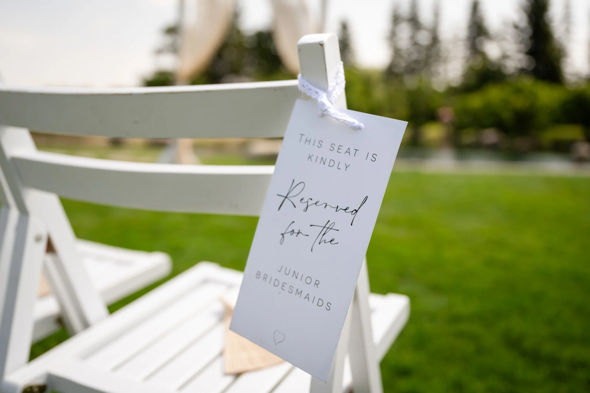 White chair with a reserved sign hanging from the backrest, outdoor green grass and trees in the background.