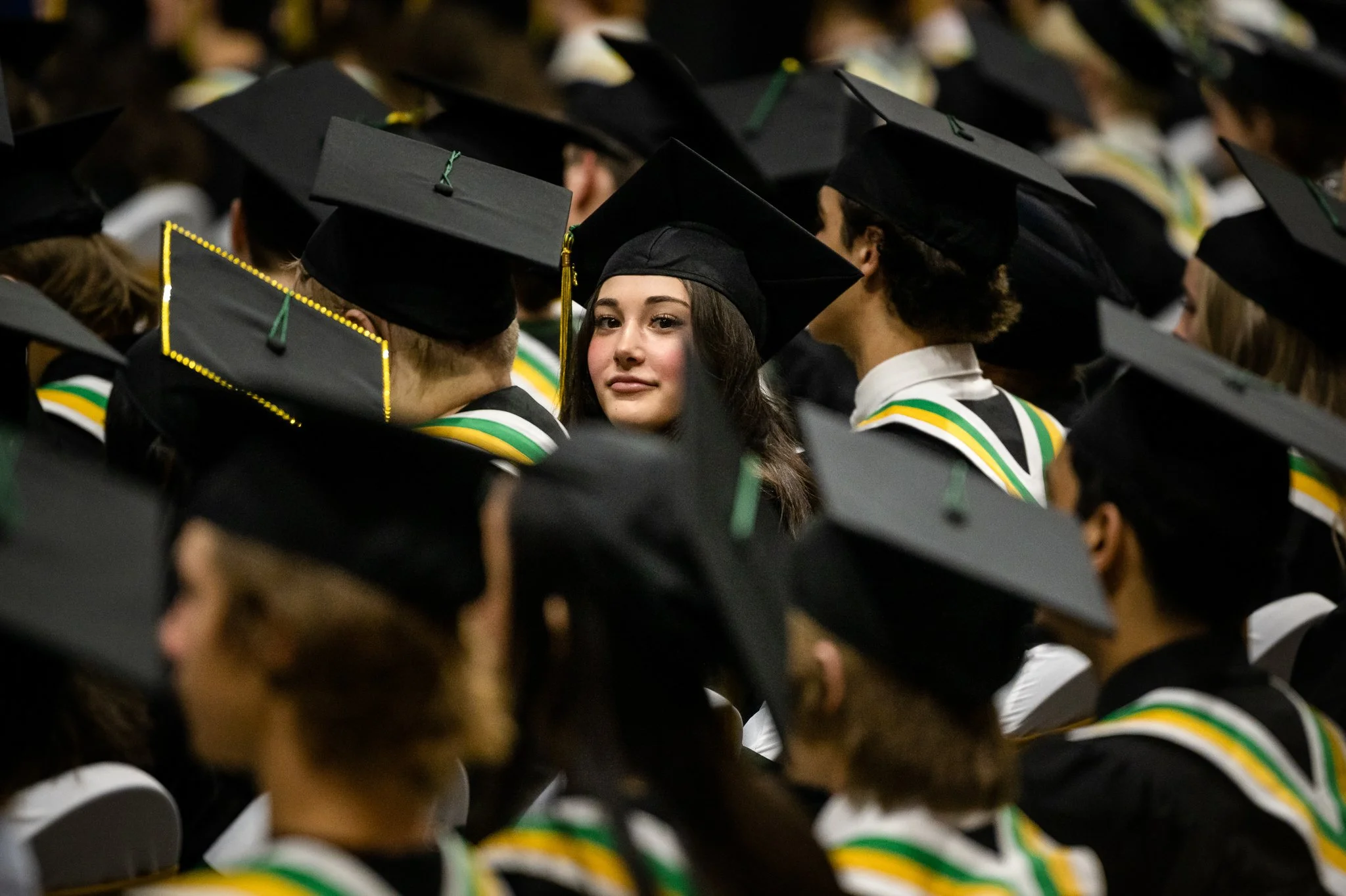 A group of graduates in black caps and gowns with green, yellow, and white stoles, gathered at a graduation ceremony, with one young woman looking directly at the camera.