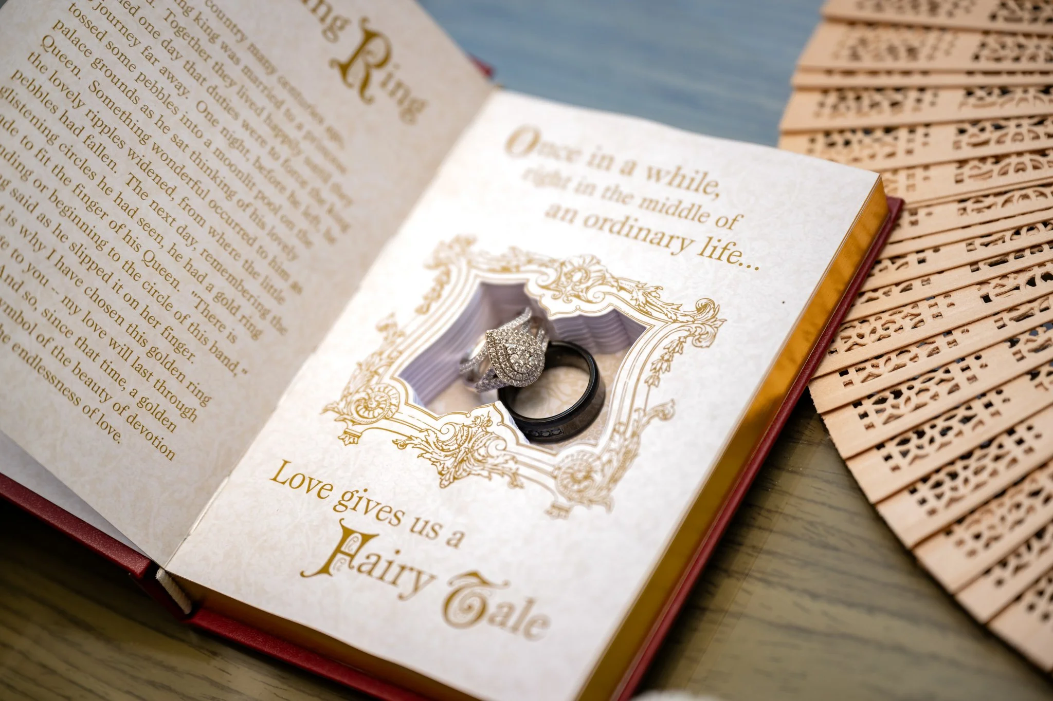 An open storybook with wedding rings placed inside a decorative frame on the page, and the text 'Once in a while, in the middle of an ordinary life... Love gives us a fairy tale.' The book rests on a wooden surface with a carved wooden fan nearby.