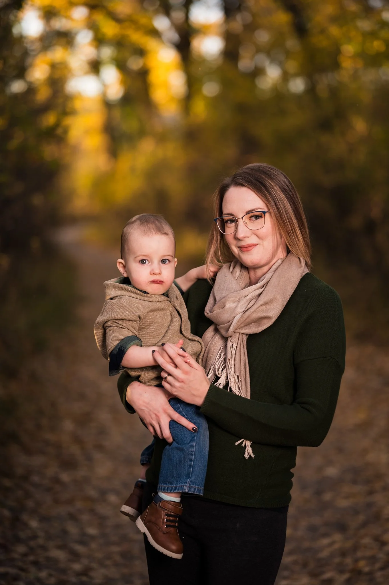 A woman with glasses and a beige scarf holding a young boy in an autumn forest.