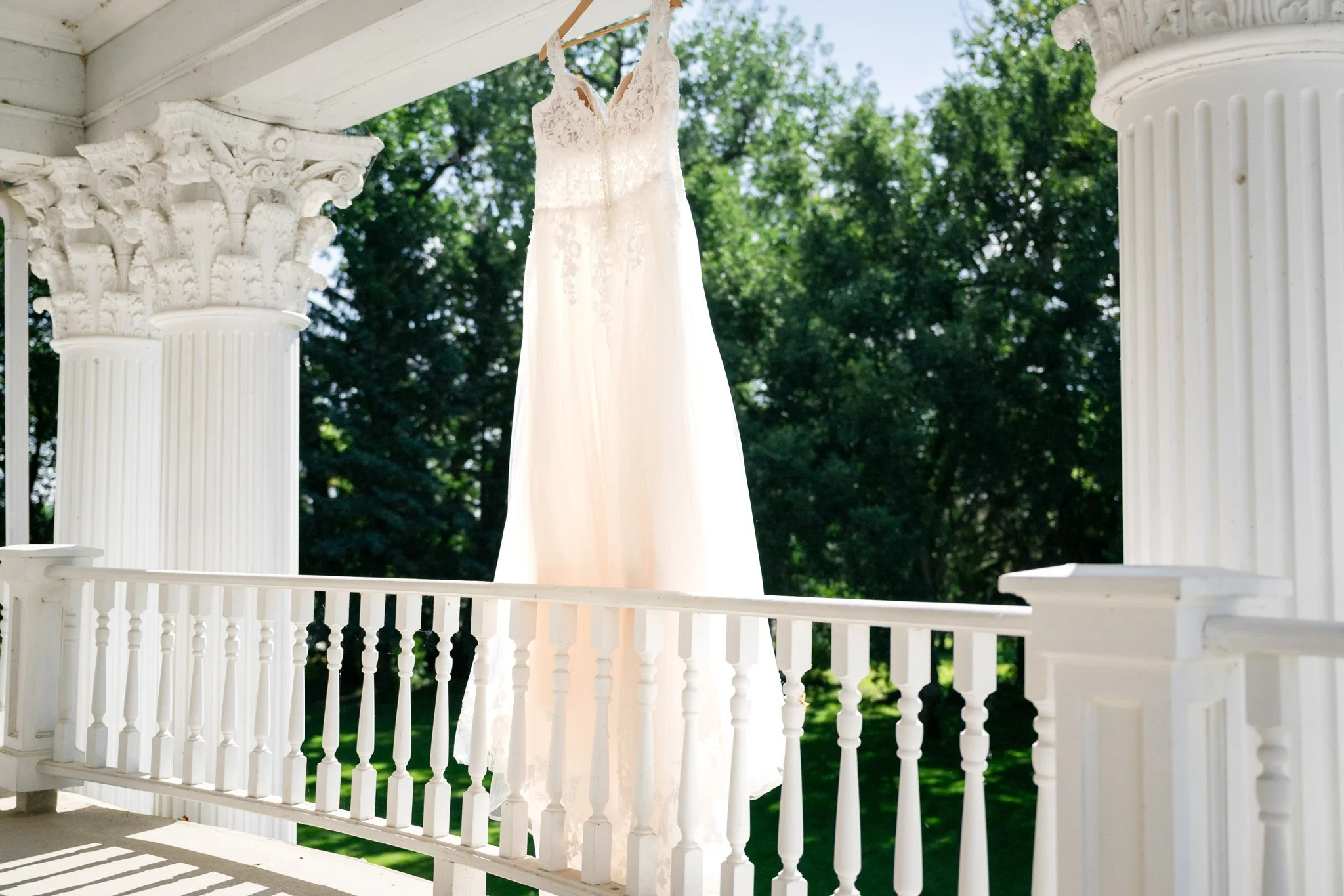 A white wedding dress hanging from a porch ceiling with white columns and railing, outdoors with green trees in the background.