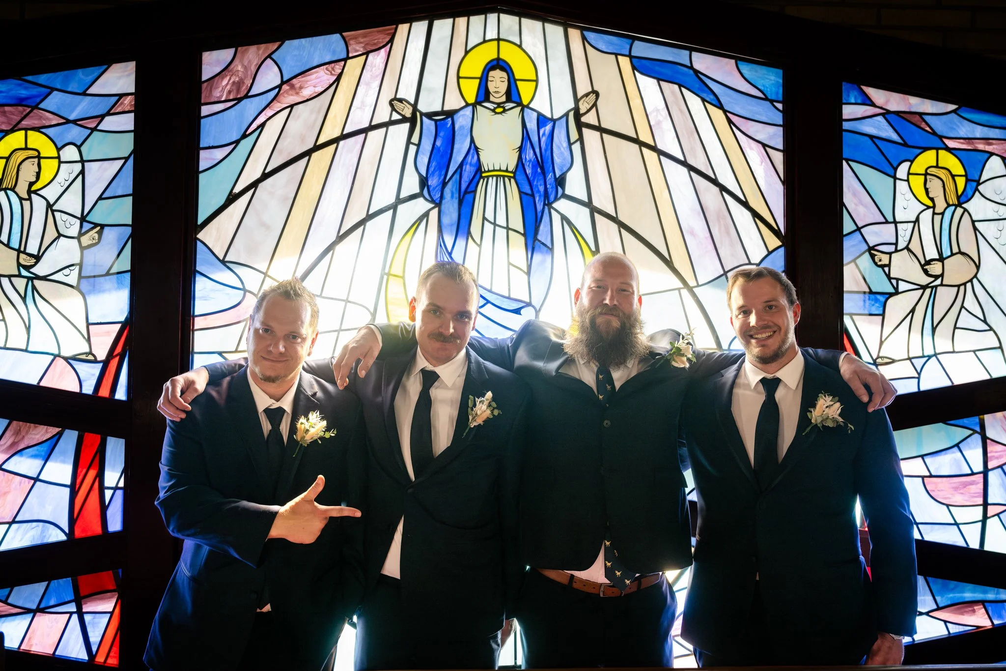 Four men in suits with boutonnières, standing arm-in-arm in front of a large stained glass window depicting Jesus with open arms and angels on either side.