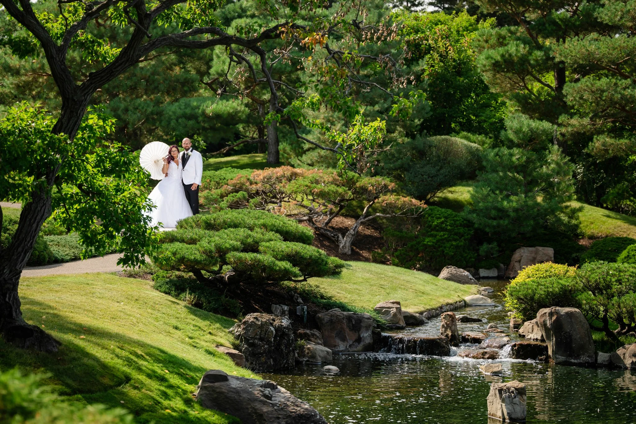A bride and groom stand together in a lush, green garden near a small waterfall, surrounded by trees and shrubs, with the bride holding a white parasol.