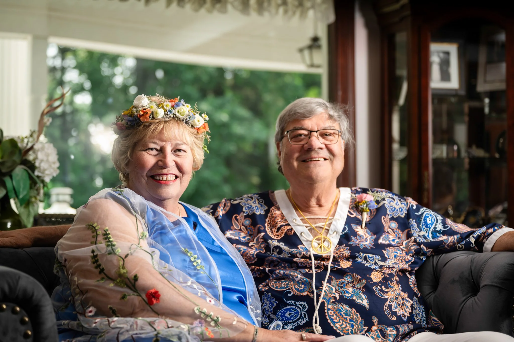 An elderly woman and man sitting on a black leather couch, smiling. The woman is wearing a flower crown and a sheer embroidered shawl, the man is wearing glasses and a colorful paisley shirt with peace sign necklace, in a cozy indoor setting with lar
