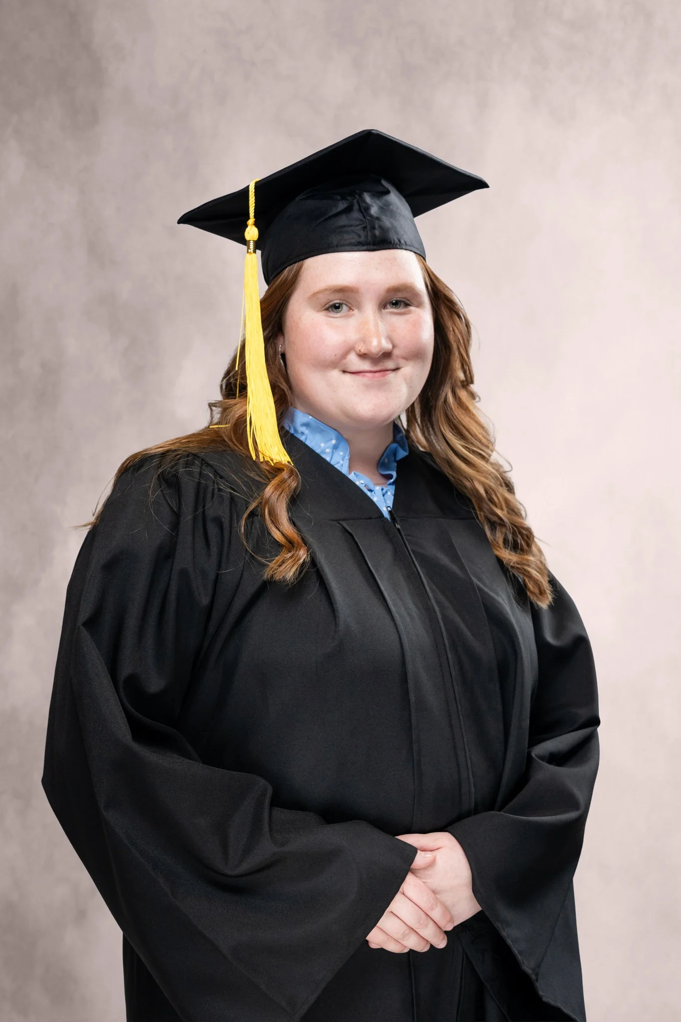Young woman in black graduation gown and cap with a yellow tassel, standing in front of a plain light background.